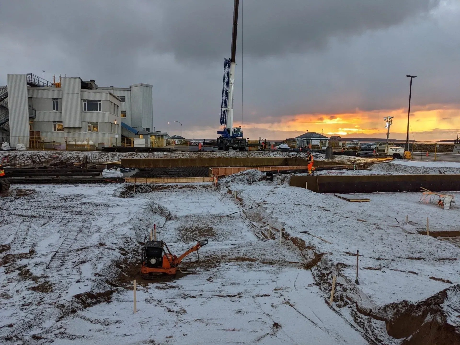 Un chantier de construction enneigé au coucher du soleil, avec une grande grue et une petite machine de compactage.
