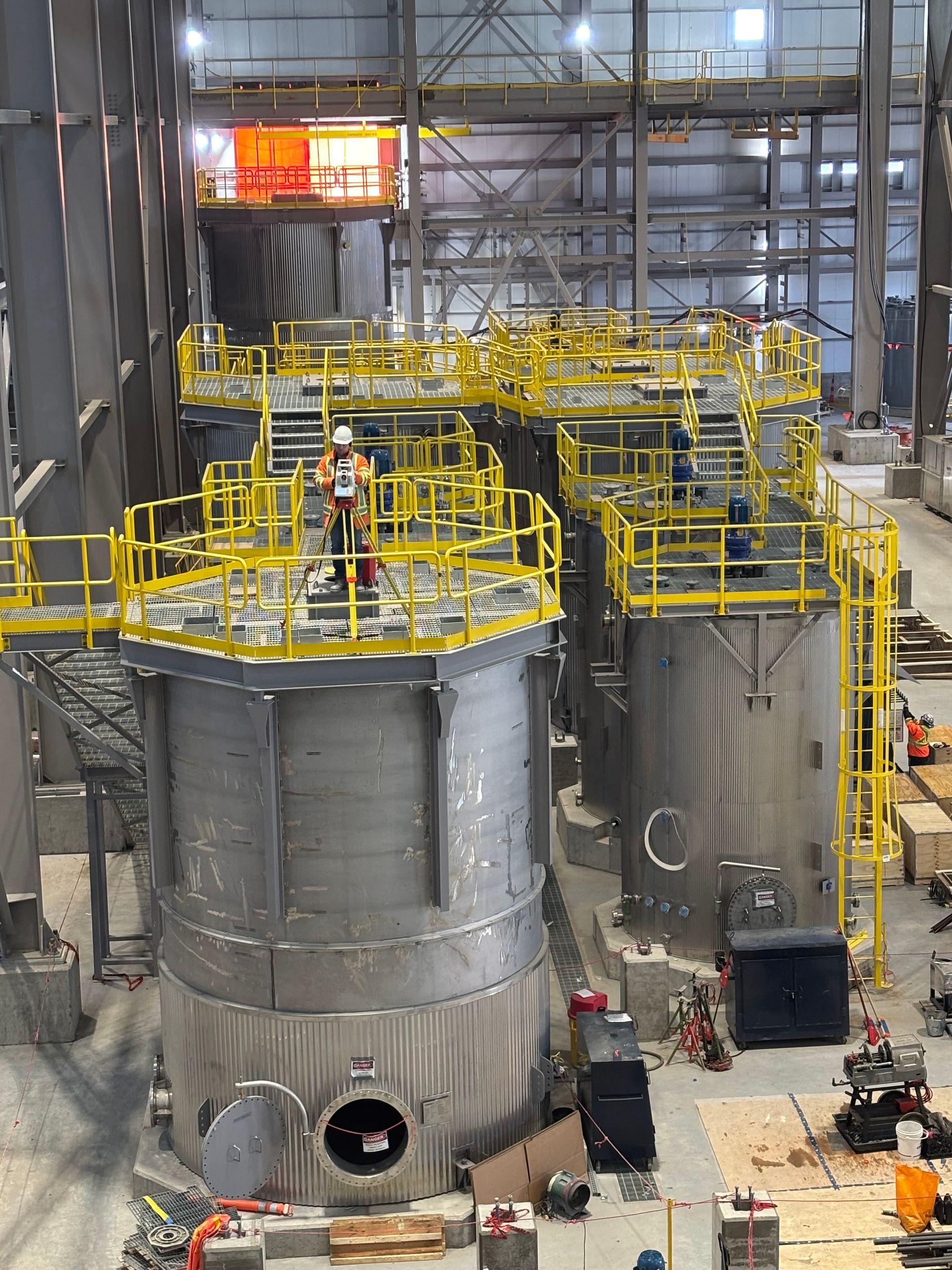 Un ouvrier du bâtiment se tient sur une plateforme métallique au-dessus de grands réservoirs industriels à l'intérieur d'un entrepôt entouré de garde-corps jaunes.