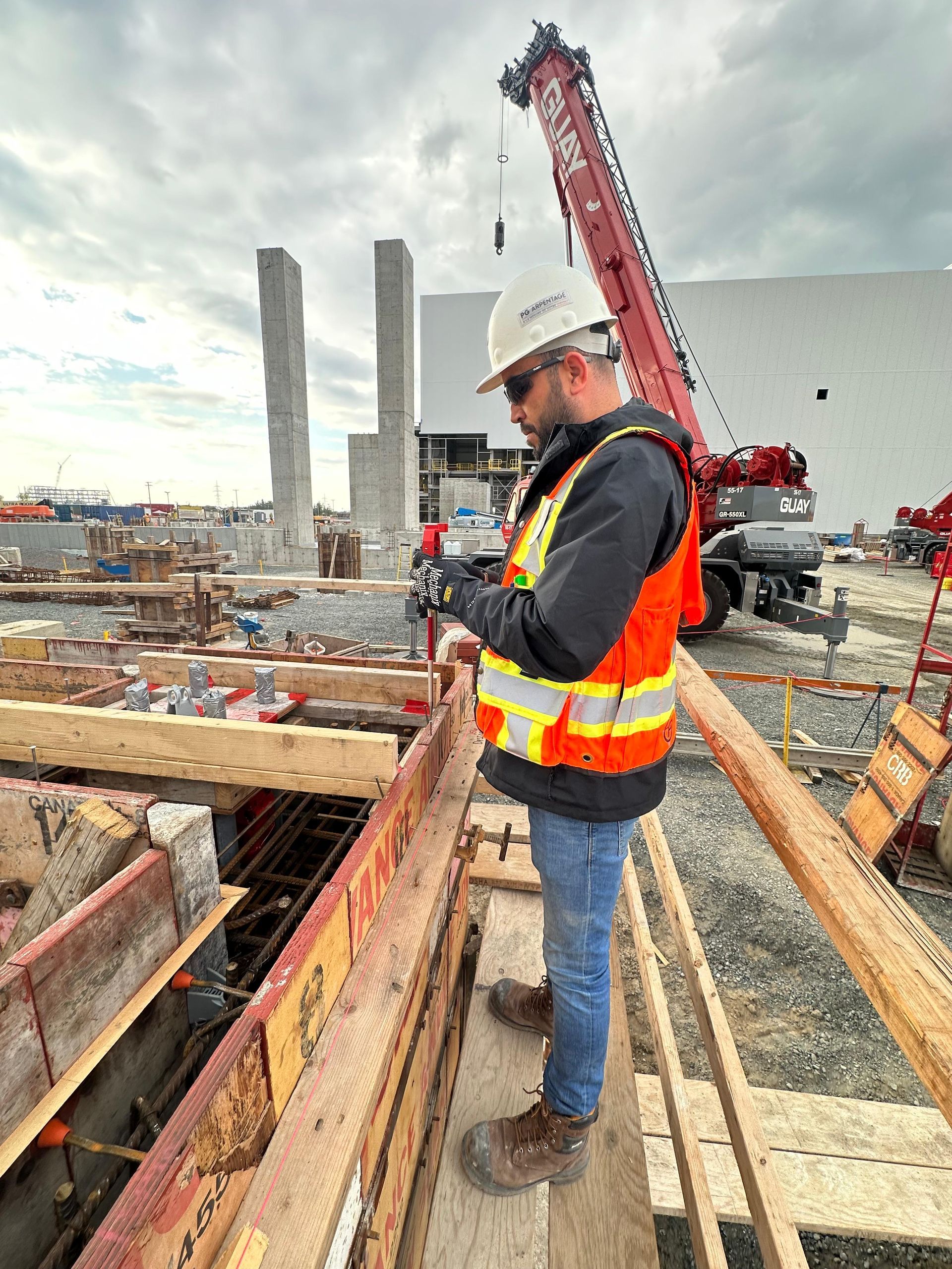 Un ouvrier du bâtiment, portant un casque et un gilet haute visibilité, examine des plans sur un chantier avec une grue en arrière-plan.
