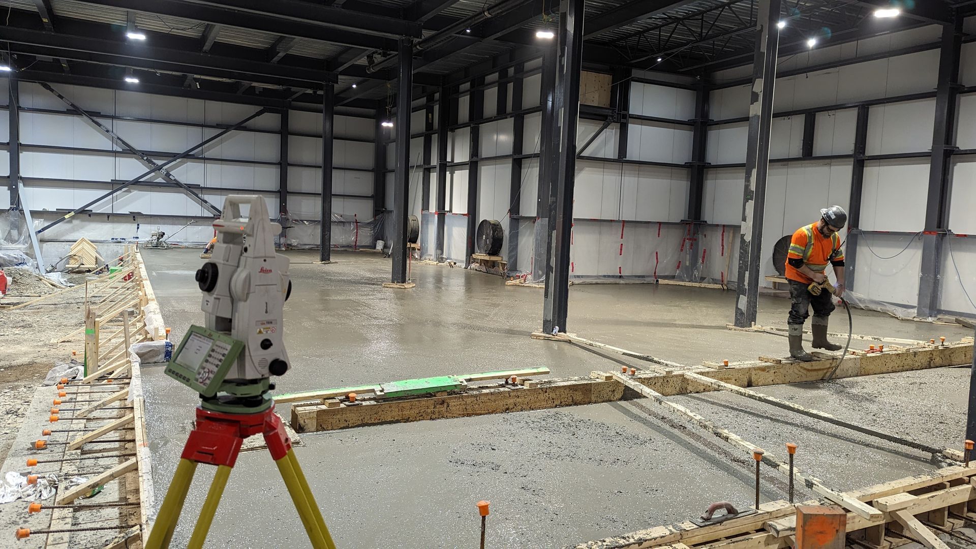 Une station totale de géomètre, montée sur un trépied, surplombe un ouvrier du bâtiment debout sur un sol en béton fraîchement coulé.