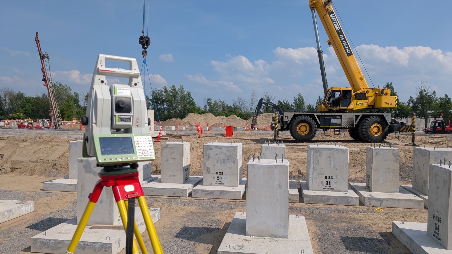 Un instrument de topographie monté sur un trépied jaune surplombe un chantier de construction avec des piliers en béton et une grue mobile jaune.