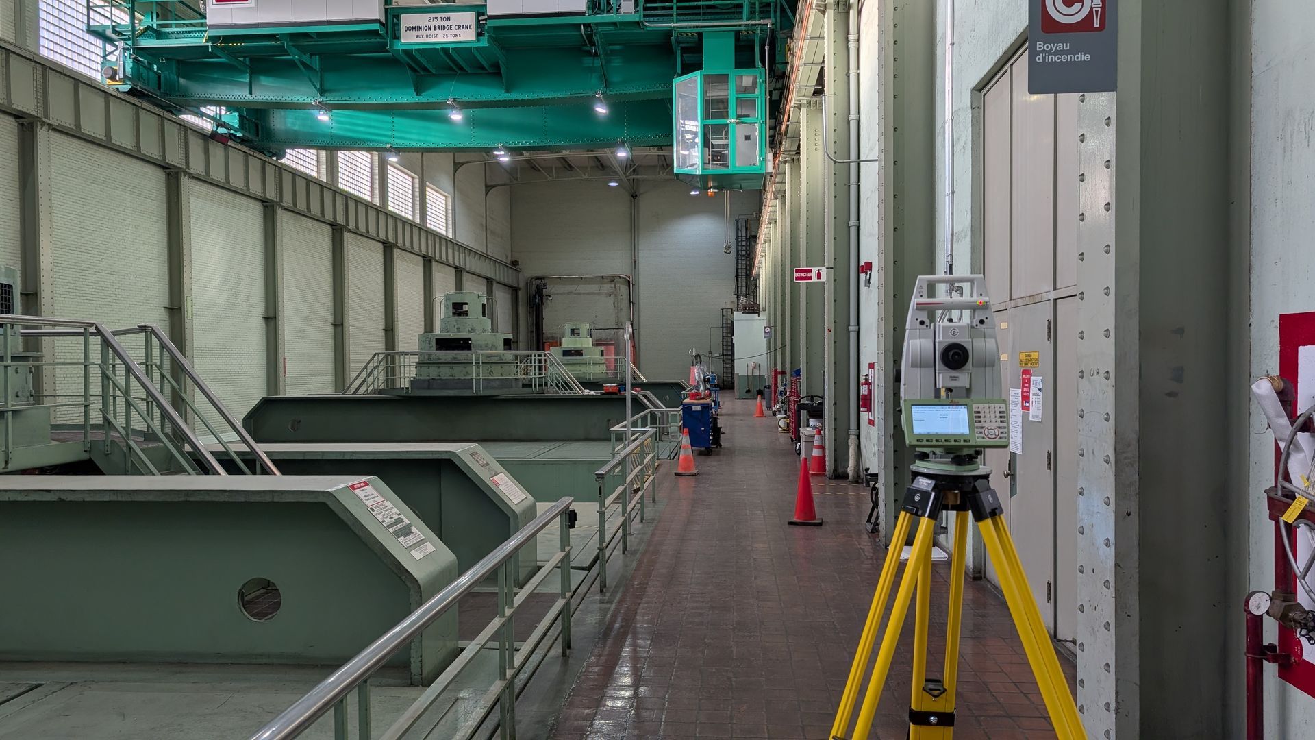 Un instrument de topographie par station totale sur un trépied jaune à l'intérieur d'une usine industrielle avec de grosses machines et une grue au-dessus.