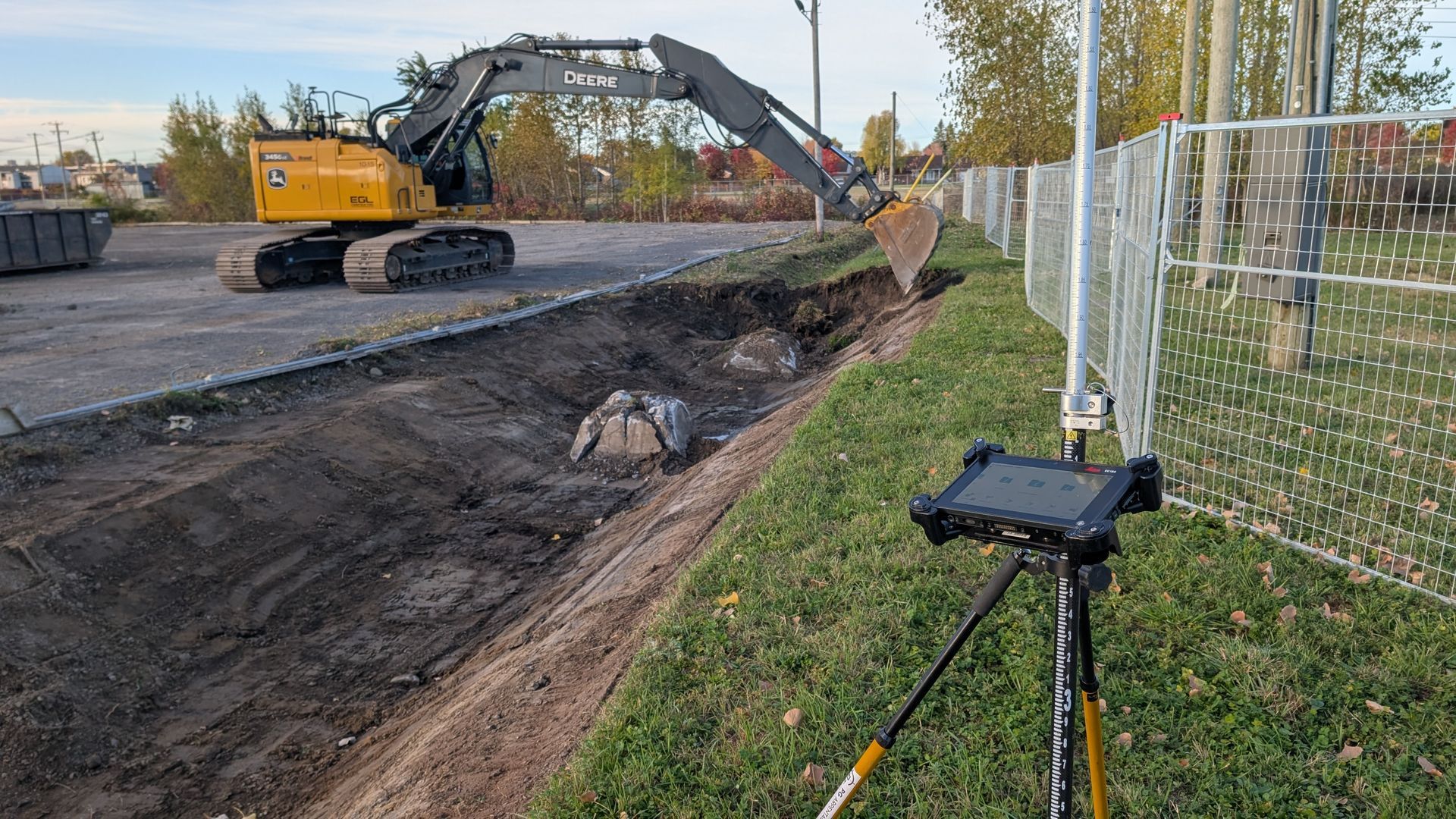 Une pelleteuse jaune creuse une tranchée sur un chantier, sous le regard d'une tablette fixée sur un trépied à proximité.
