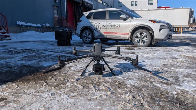 Un drone est posé sur un sol enneigé devant un SUV blanc arborant un logo, garé près d'un bâtiment.