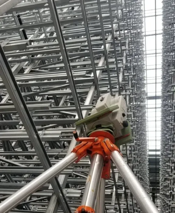 Un niveau optique de géomètre sur un trépied dans un grand entrepôt rempli de rangées d'étagères métalliques.