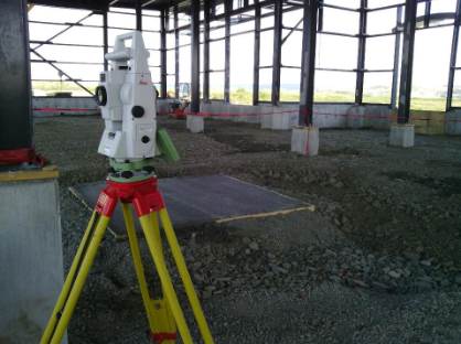 Une station totale de topographie sur un trépied jaune à l'intérieur d'un bâtiment industriel à ossature métallique en construction.