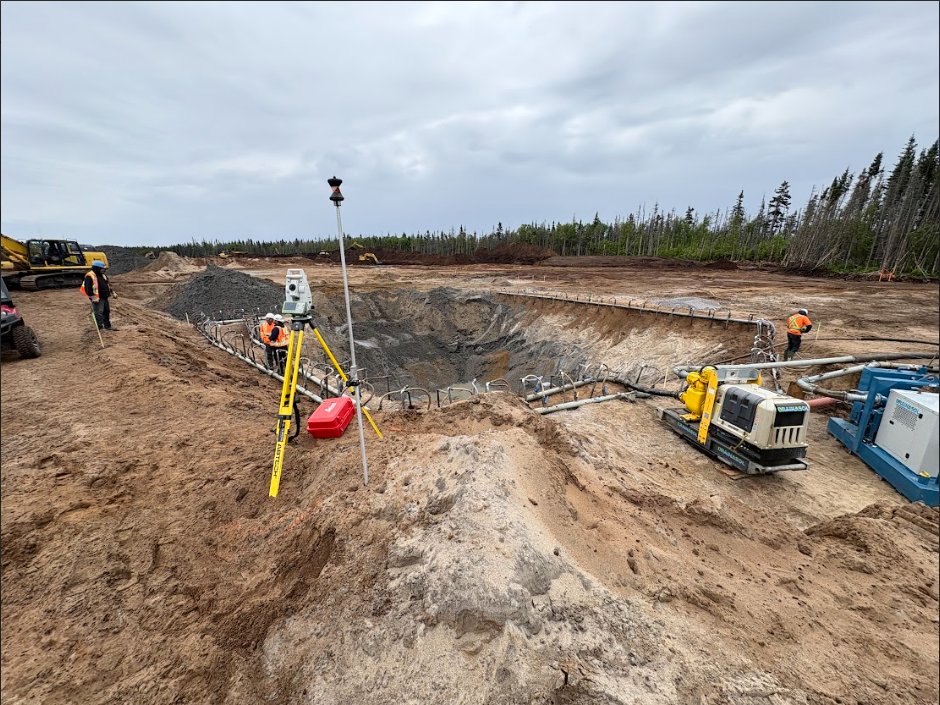 Des ouvriers et du matériel topographique sur un grand chantier de construction excavé, avec une forêt en arrière-plan.