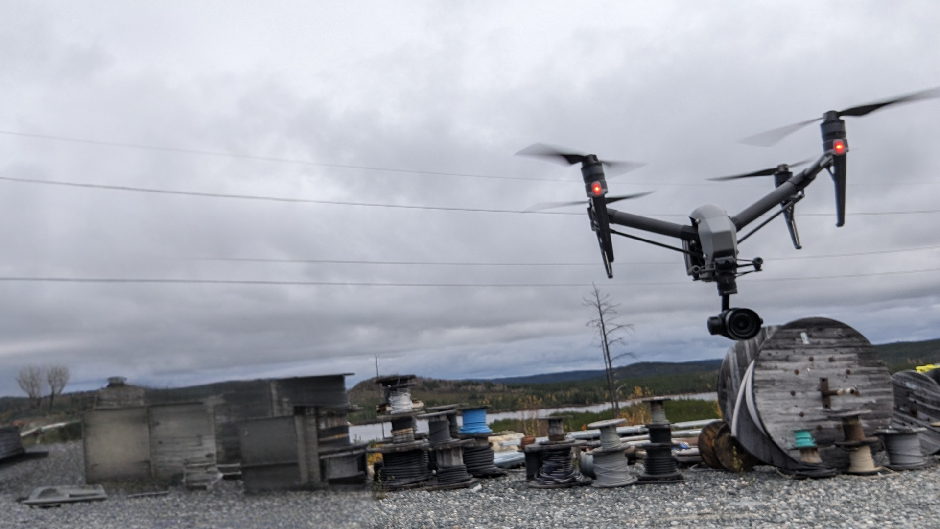 Un drone gris survole un champ parsemé de bobines et d'équipements industriels divers, sous un ciel nuageux.