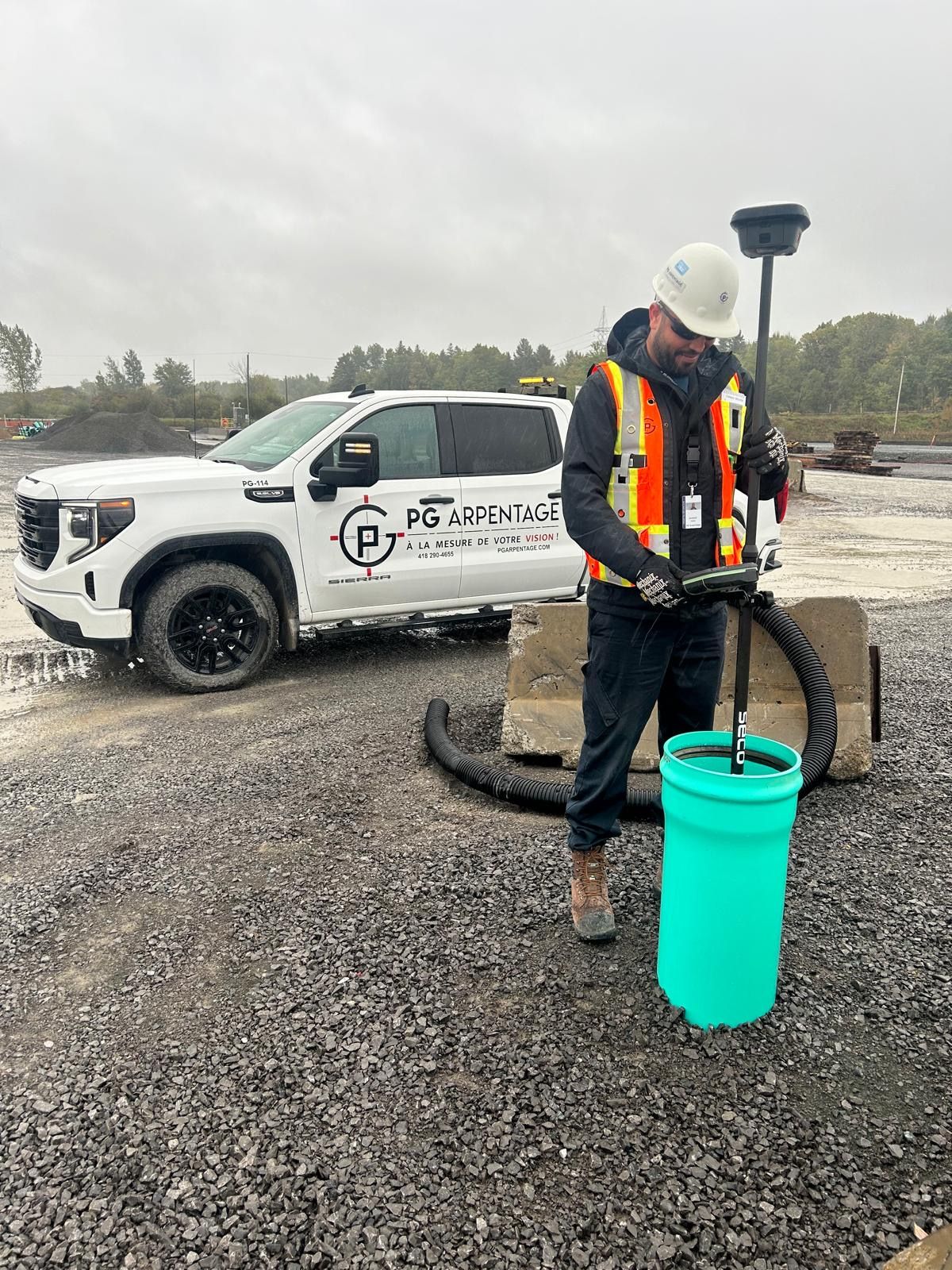 Un ouvrier portant un gilet haute visibilité utilise une perche d'arpenteur pour mesurer un seau en plastique turquoise sur un chantier de construction.