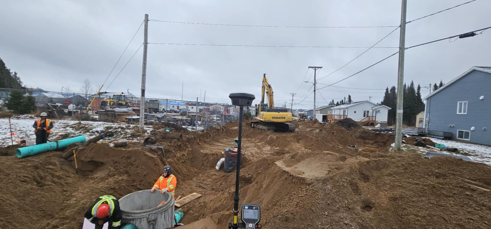 Des ouvriers du bâtiment, équipés de tenues de sécurité, installent des canalisations dans une tranchée creusée, tandis qu'une excavatrice travaille en arrière-plan.