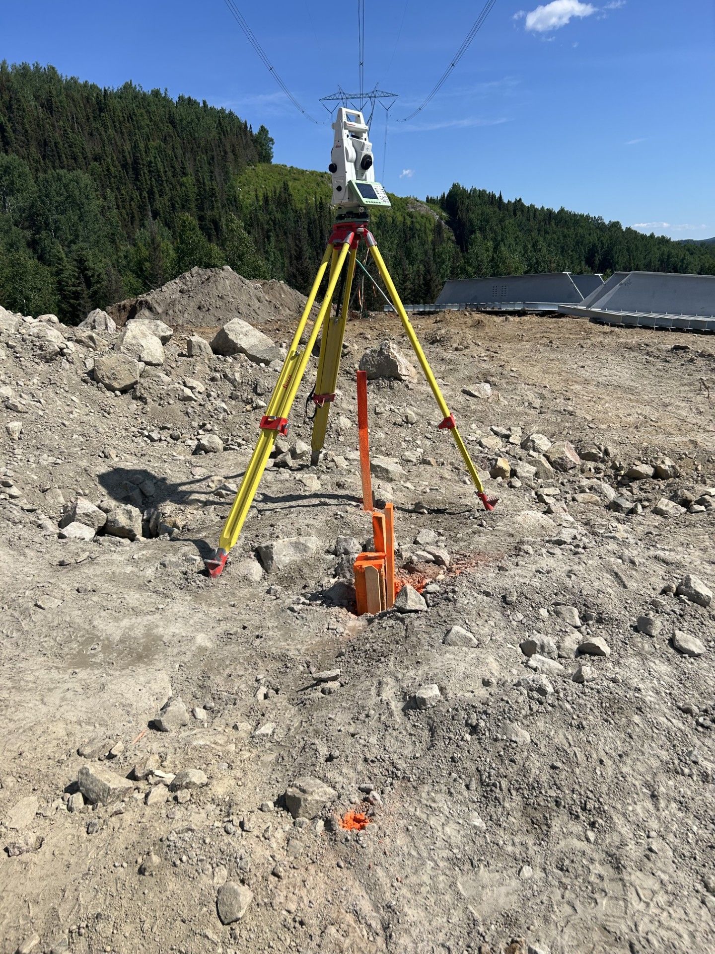 Une station totale de topographie, montée sur un trépied jaune, se dresse sur un terrain rocailleux dans une zone de construction, sous un ciel bleu dégagé.