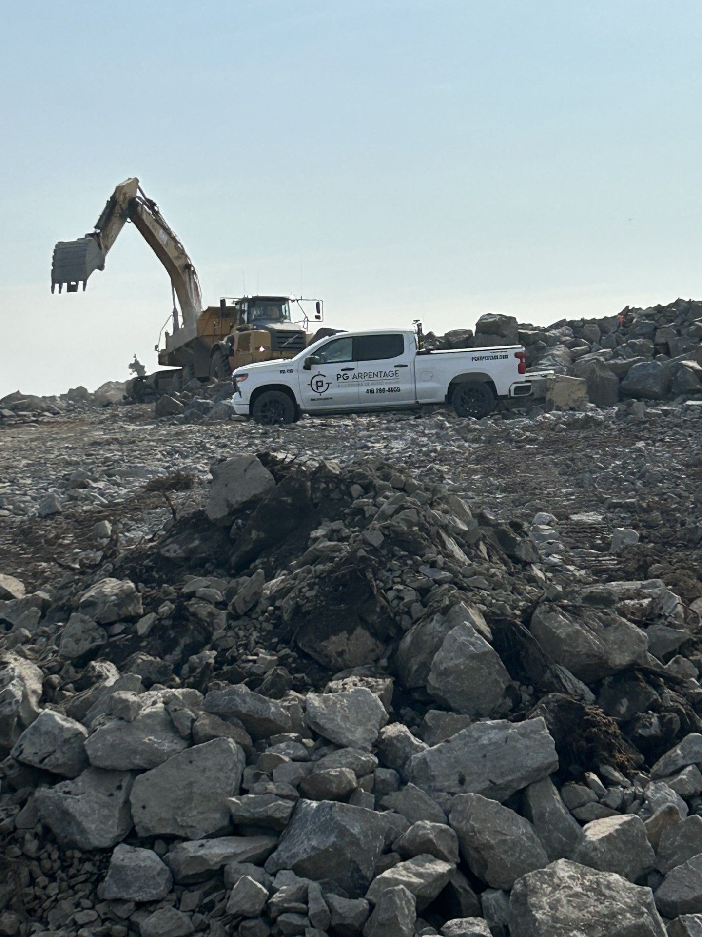 Une pelleteuse jaune et une camionnette blanche sur un terrain de démolition rocailleux et accidenté, sous un ciel clair et lumineux.