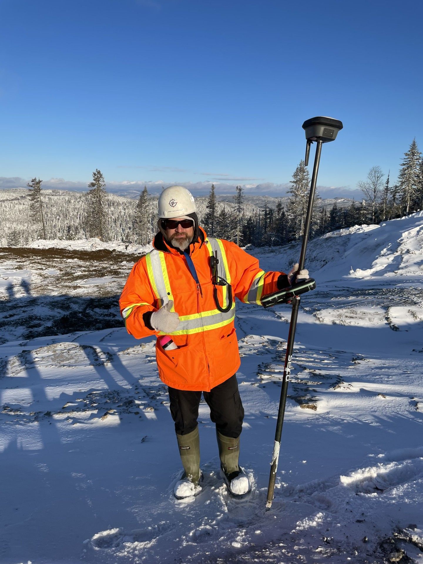 Un géomètre, vêtu d'une veste orange haute visibilité et d'un casque de chantier, se tient debout dans un paysage enneigé, tenant une perche d'arpenteur.