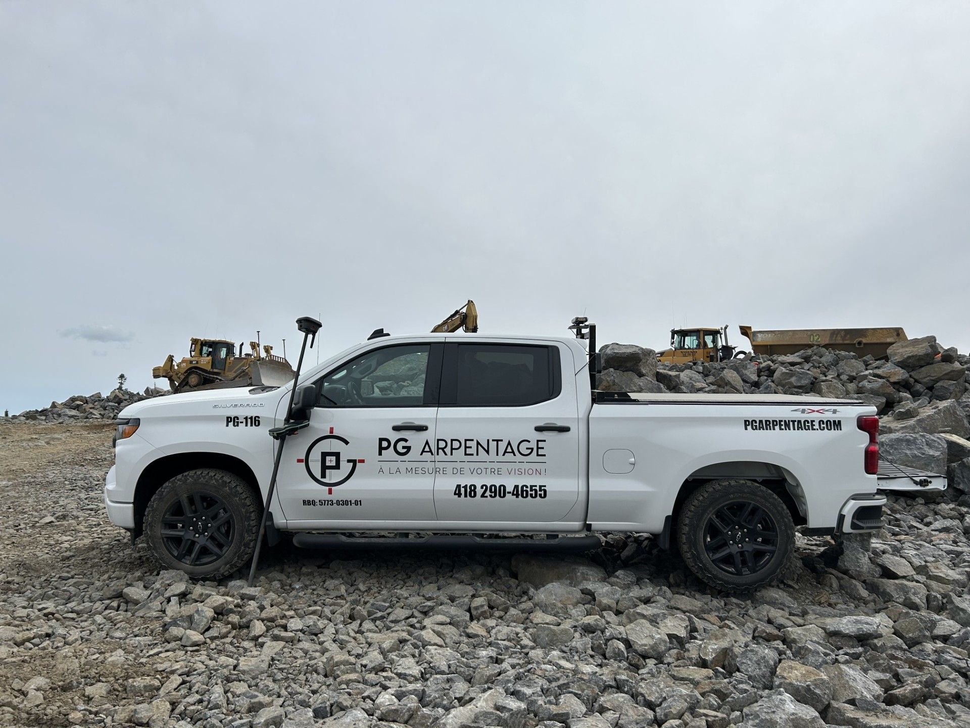 Un camion d'arpentage blanc est stationné sur un chantier rocailleux, avec de la machinerie lourde en arrière-plan.