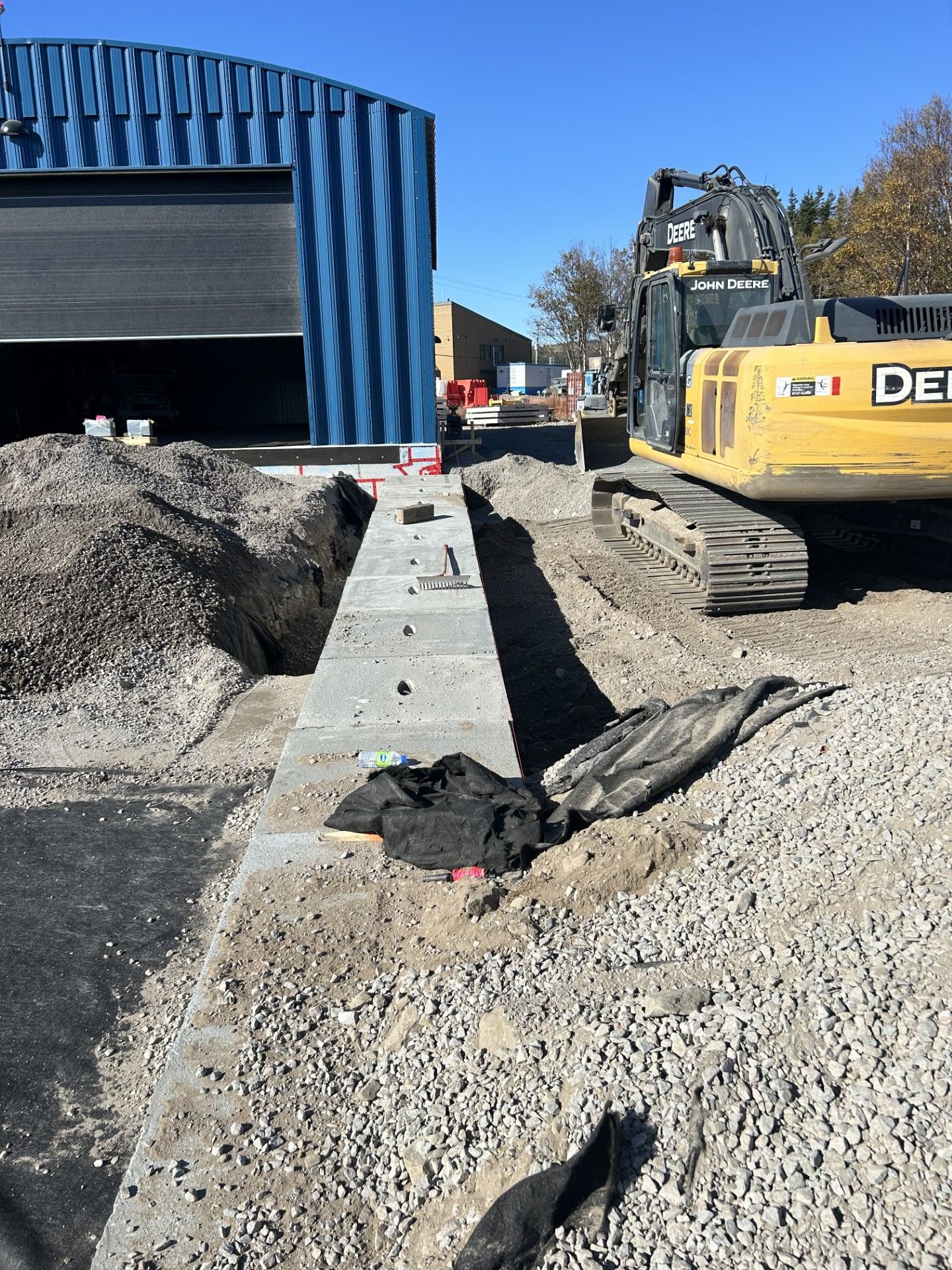 Une pelle mécanique Deere jaune est garée à côté d'un mur de fondation en béton fraîchement coulé, devant un bâtiment industriel bleu.