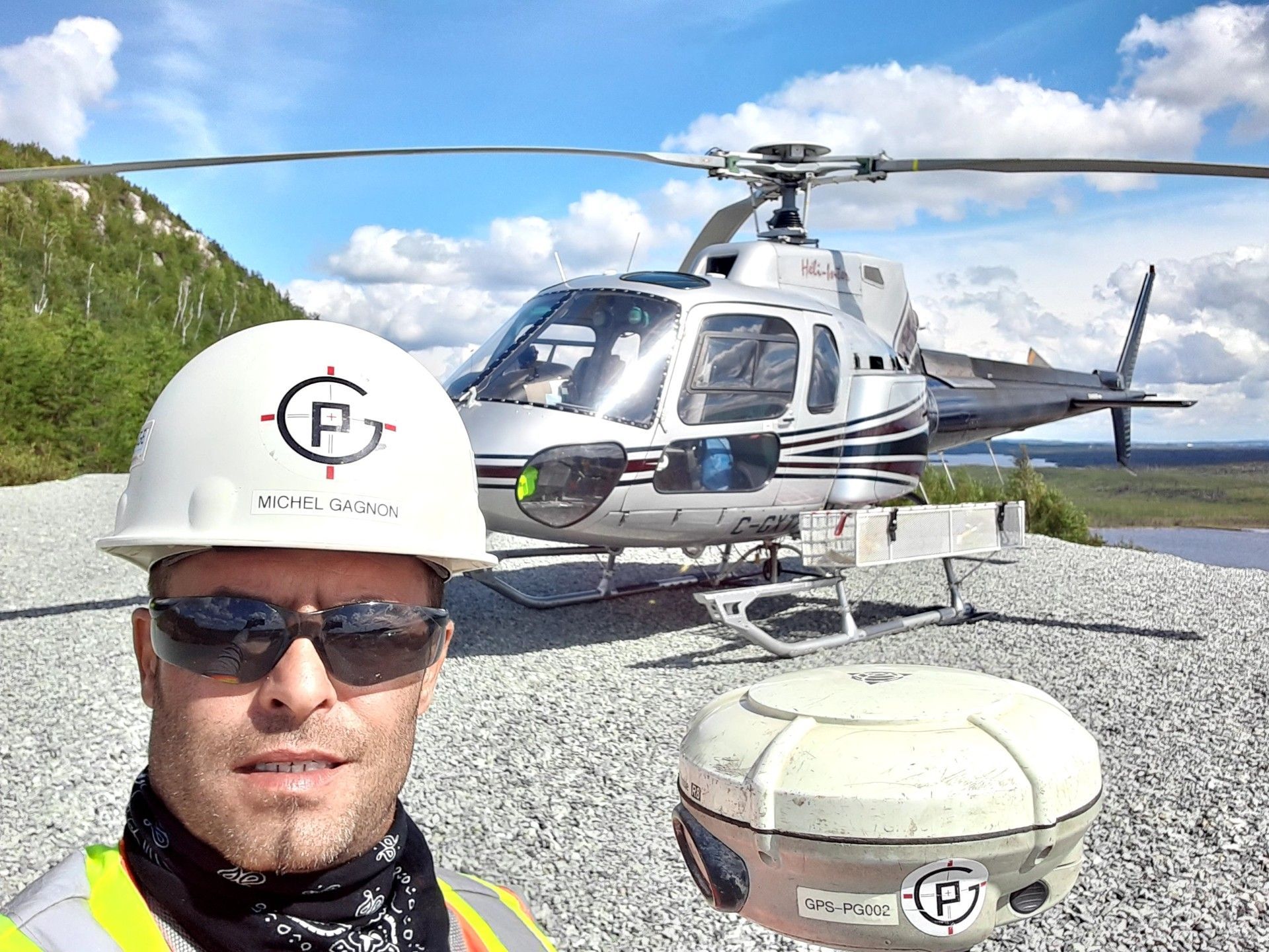 Un professionnel portant un casque de chantier blanc orné d'un logo prend un selfie à côté d'un hélicoptère argenté sur un héliport rocailleux.