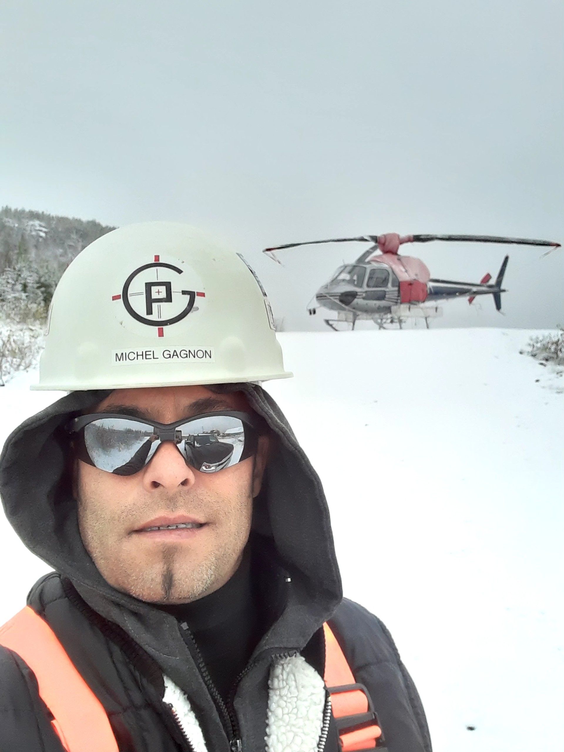 Un homme portant un casque de chantier, un sweat à capuche et des lunettes de soleil dans un champ enneigé, avec un hélicoptère en arrière-plan.