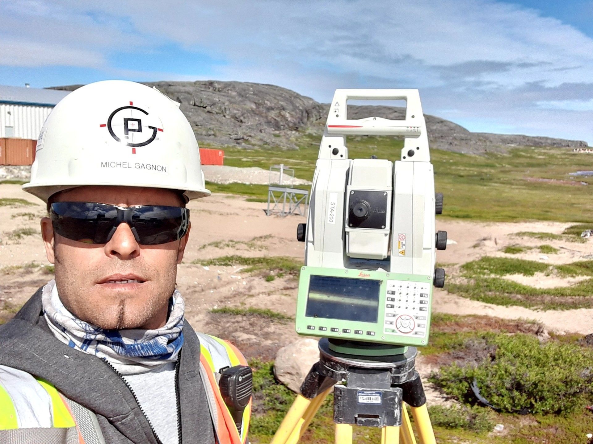 Un géomètre, portant un casque et des lunettes de soleil, prend un selfie à côté d'une station totale sur un trépied, dans un paysage rocheux.