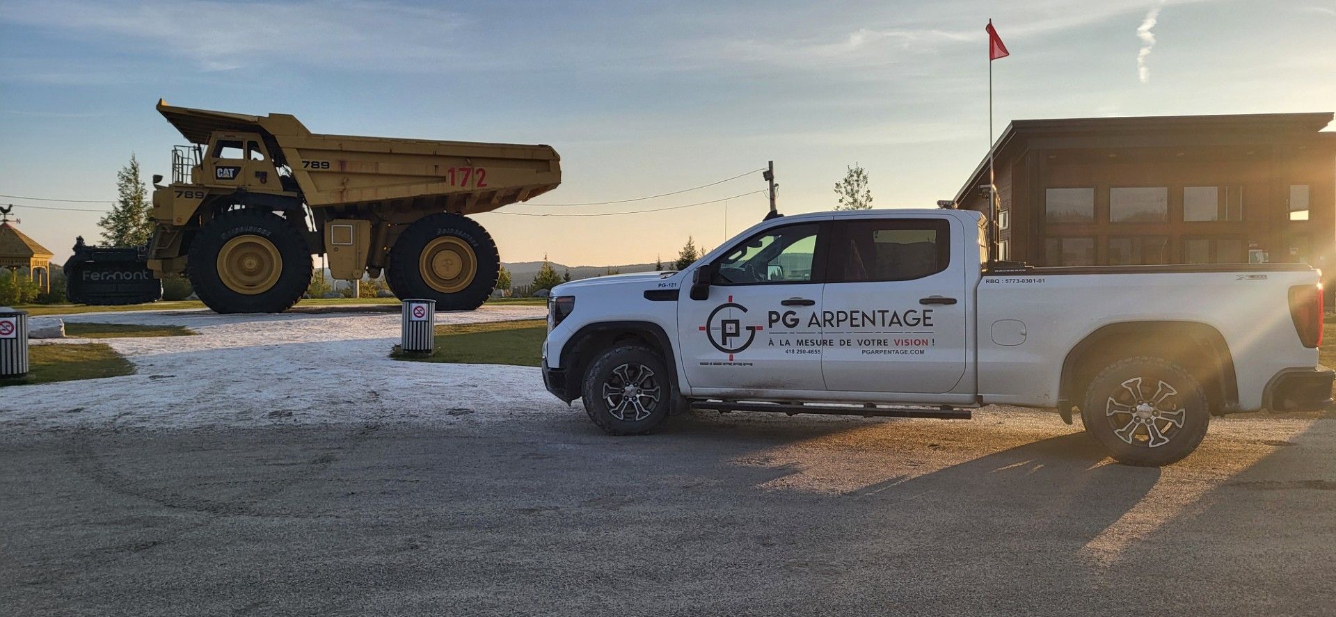 Au coucher du soleil, une camionnette blanche arborant un logo est garée sur un terrain gravillonné à côté d'un gros camion-benne jaune transportant des déchets miniers.