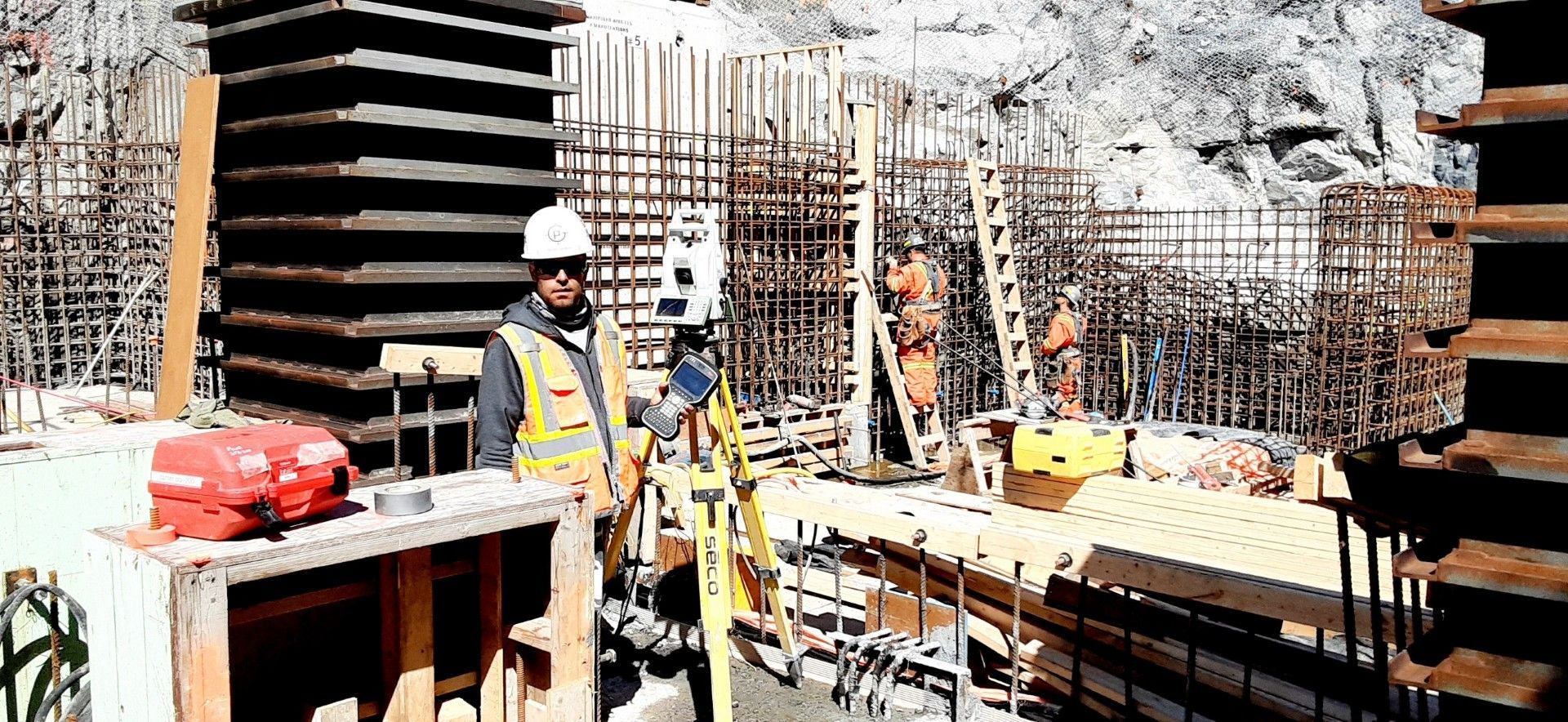 Un géomètre, portant un casque et un gilet de sécurité, utilise une station totale sur un chantier rocailleux.