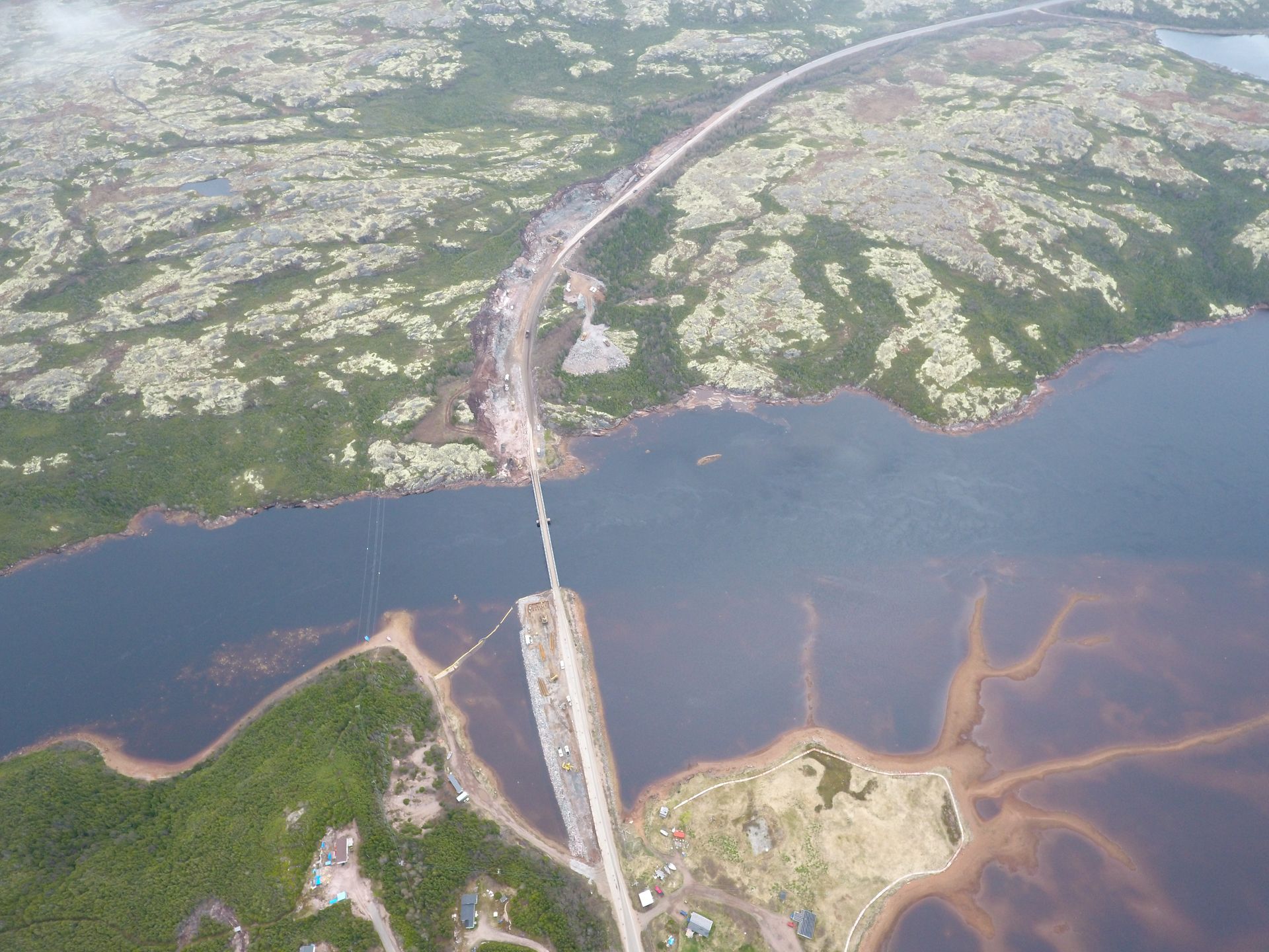 Vue aérienne d'une longue route et d'un pont traversant une étendue d'eau sombre entourée d'un terrain rocheux et verdoyant.