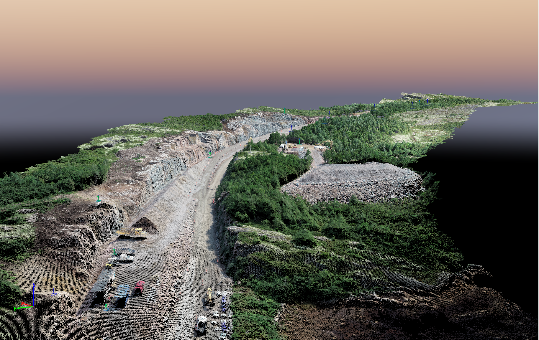 Une image aérienne 3D montre un site minier rocheux ou une tranchée d'excavation entourée d'une forêt verdoyante dense sous un ciel de coucher de soleil.