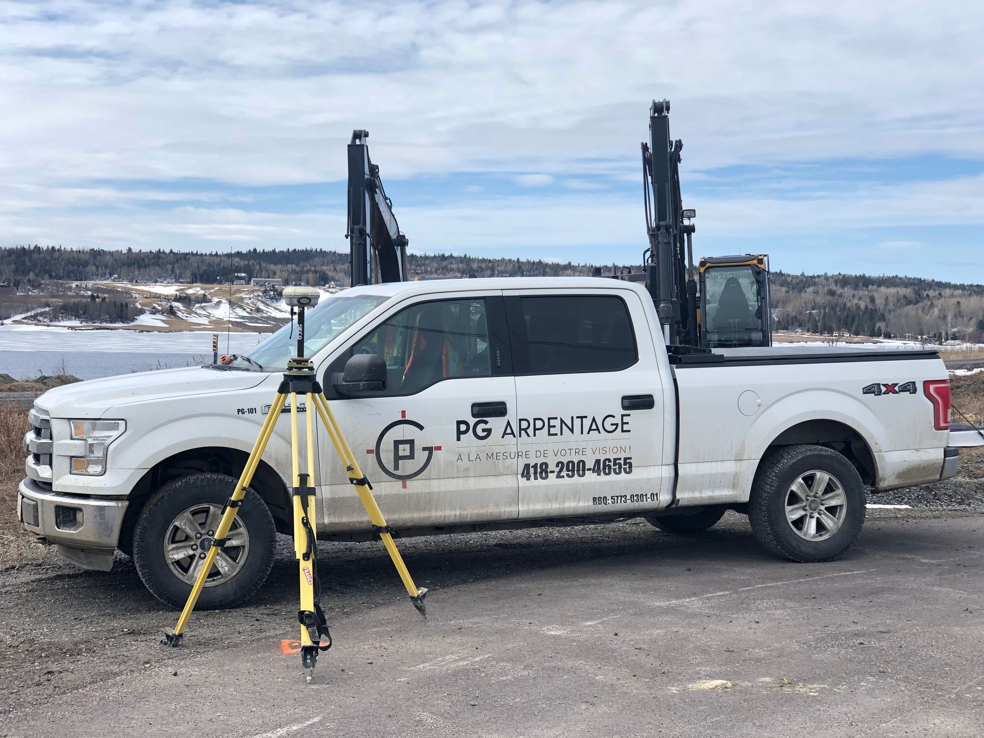 Un pick-up Ford blanc portant l'inscription « PG Arpentage » est garé à l'extérieur, avec un trépied d'arpenteur installé au premier plan.