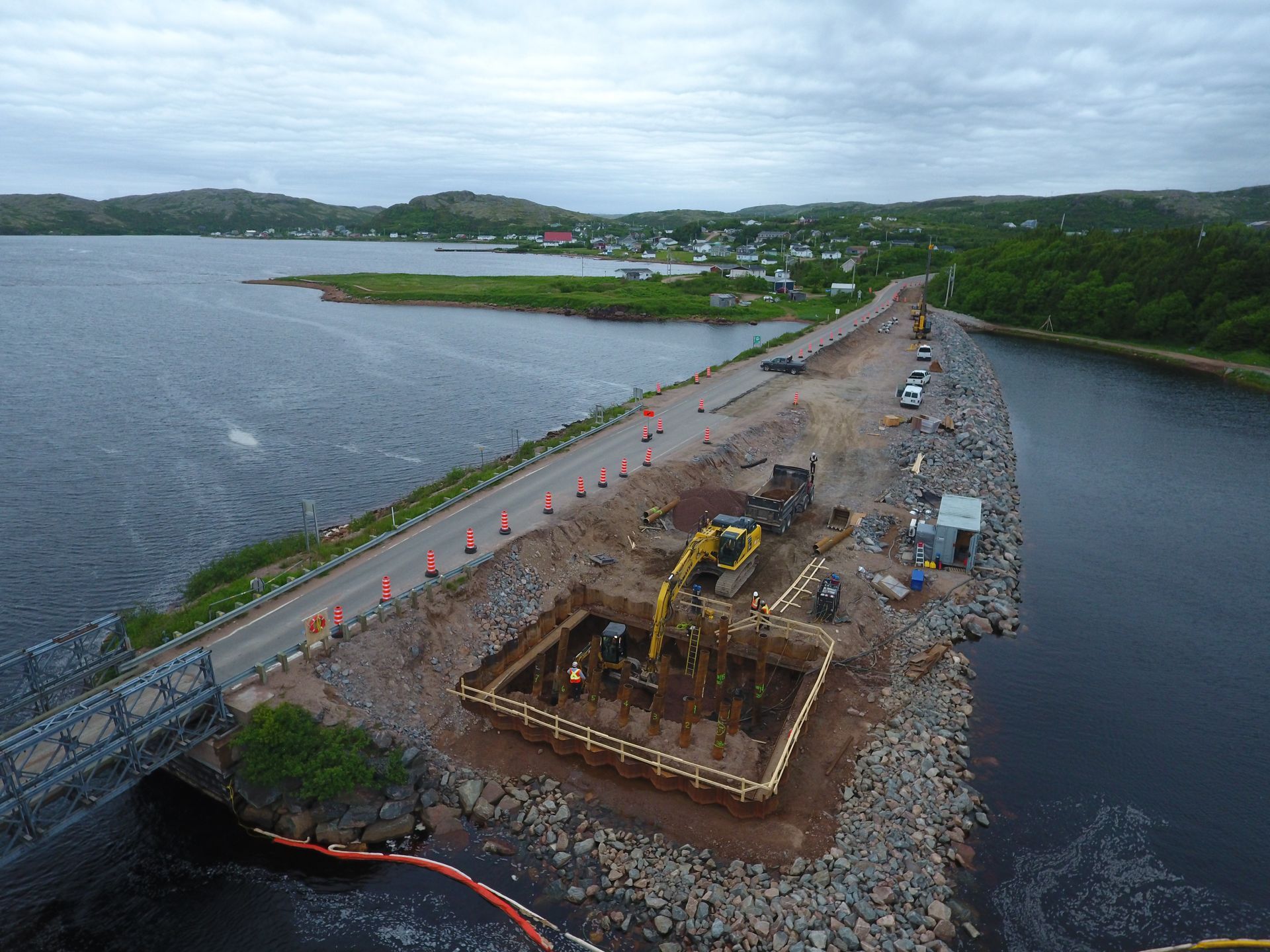 Vue aérienne d'un chantier de construction sur une chaussée reliant deux étendues d'eau, montrant une excavatrice et une fosse d'excavation.