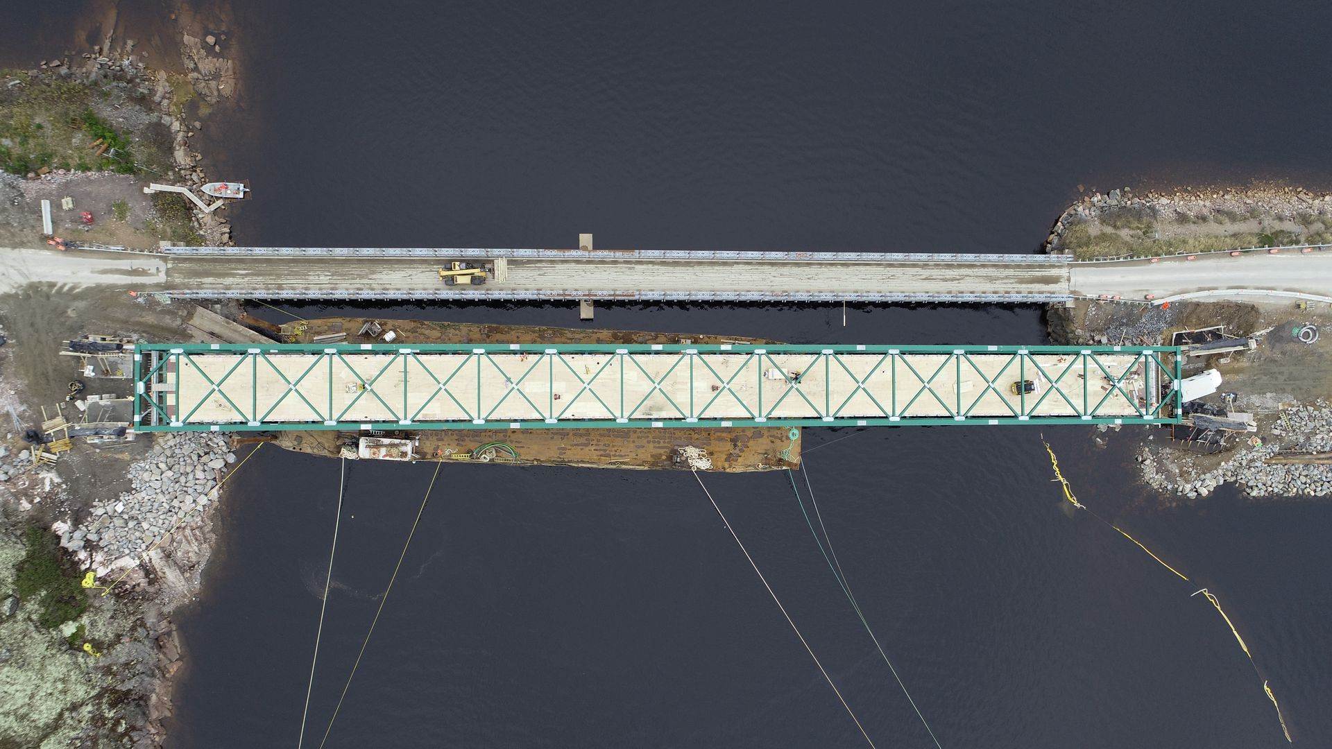 Vue aérienne d'un pont à treillis vert, parallèle à un pont routier étroit enjambant une étendue d'eau sombre.