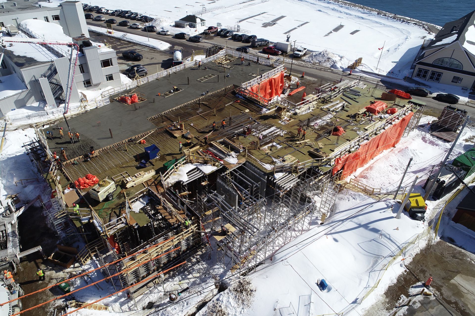 Vue aérienne d'un chantier de construction rectangulaire entouré d'un sol enneigé et d'un parking.