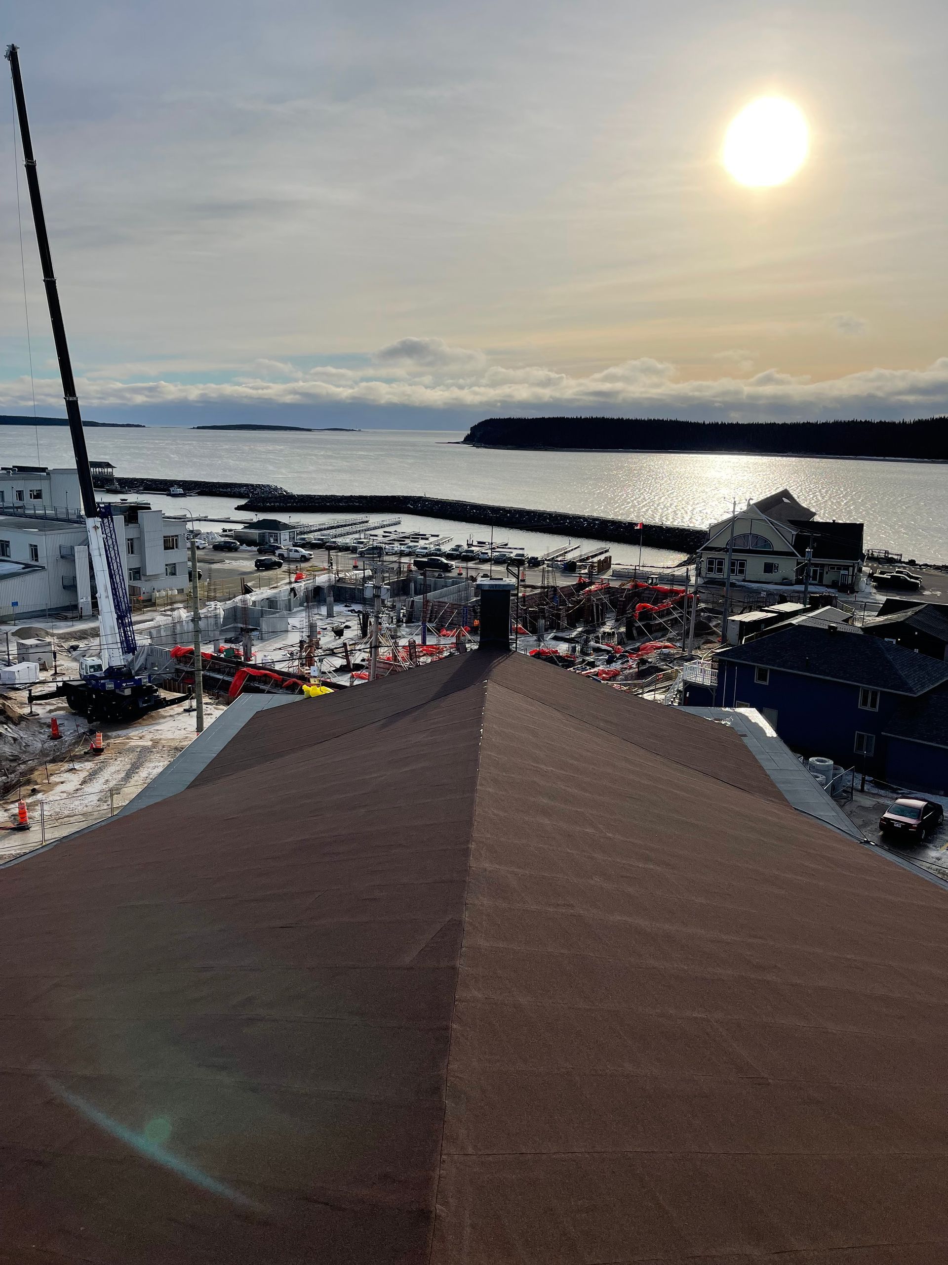 Vue en plongée d'un paysage côtier avec un toit sombre au premier plan, en direction d'un port au coucher du soleil.