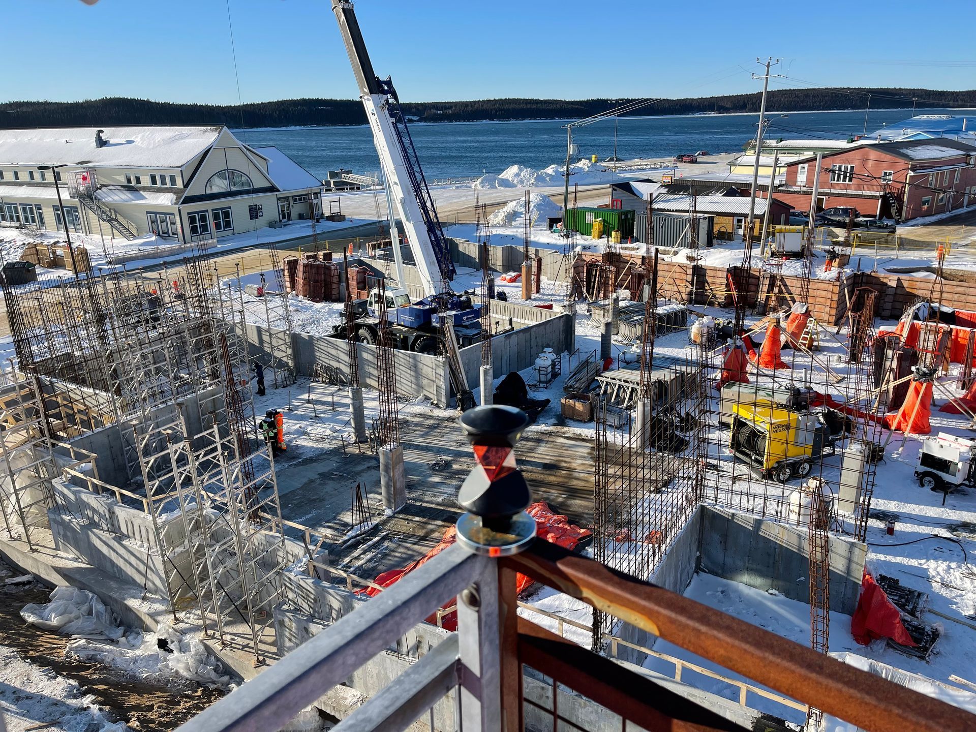 Vue en plongée d'un chantier de construction recouvert de neige, montrant une grue, une charpente métallique et des bâtiments à proximité.