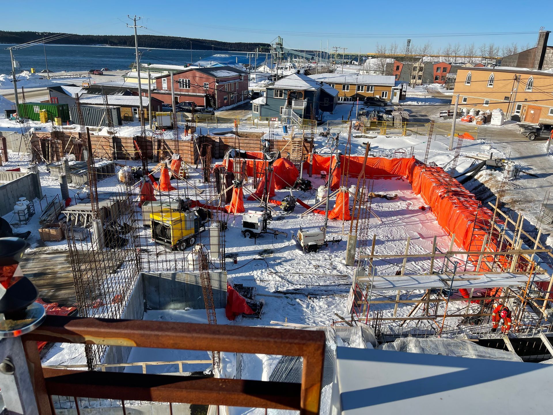 Un chantier de construction avec des ouvriers, des bâches de sécurité orange et des engins de chantier dans une zone enneigée près d'un point d'eau.