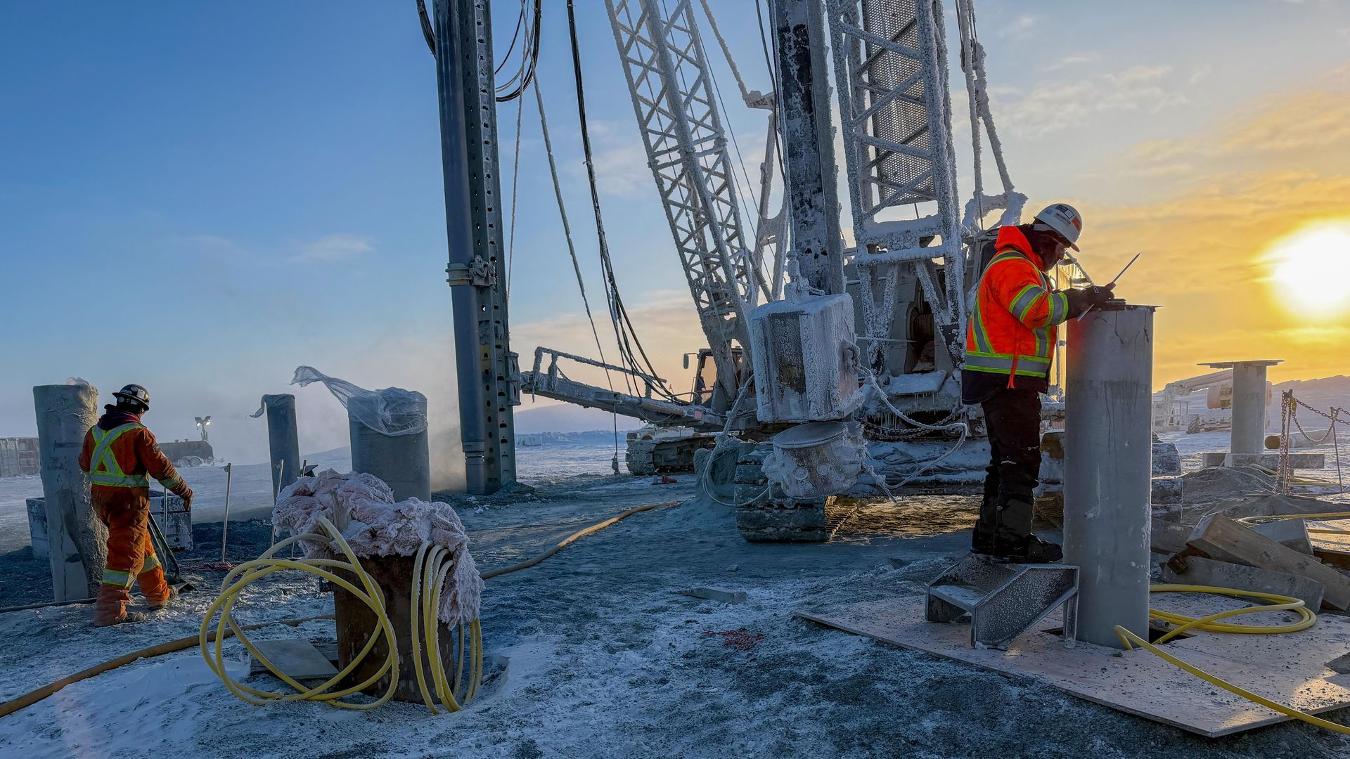 Des ouvriers portant des équipements de sécurité à haute visibilité manœuvrent de lourdes machines de forage sur un chantier enneigé et ensoleillé.