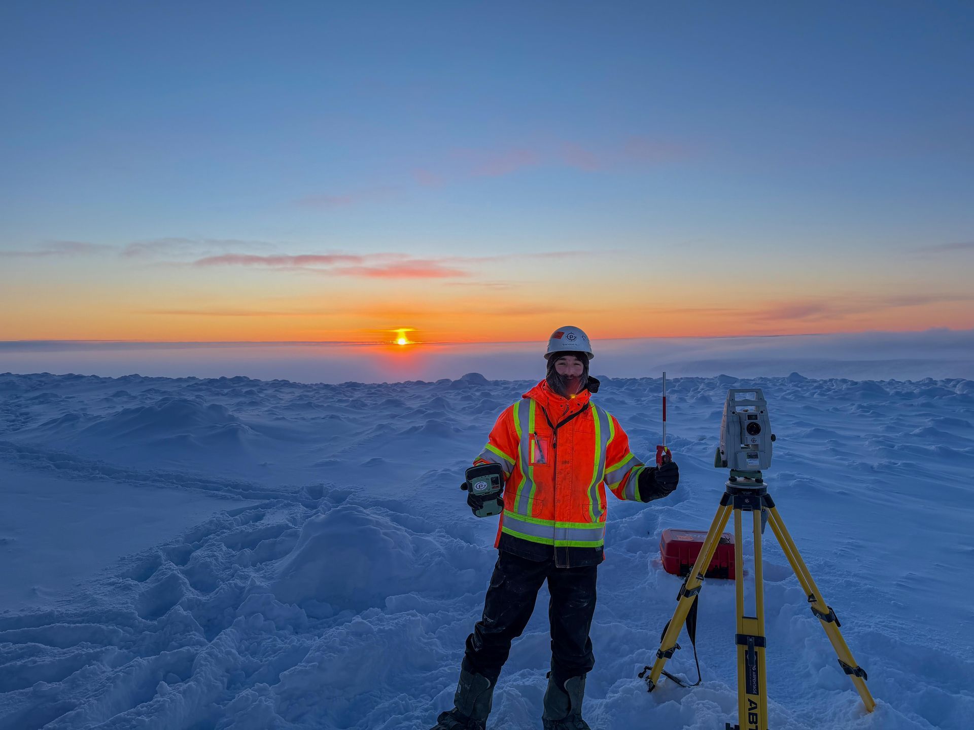 Un géomètre en tenue haute visibilité se tient sur un paysage enneigé à côté d'un instrument monté sur trépied au coucher du soleil.
