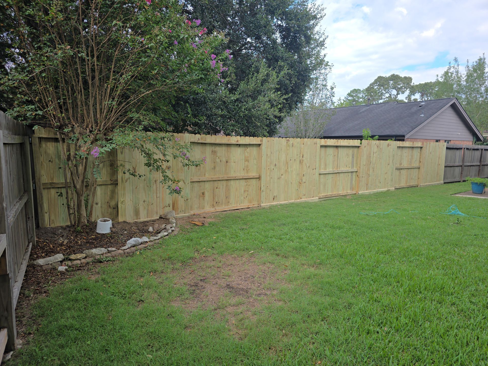 Wooden fence in a backyard with green grass and trees, under a cloudy sky.