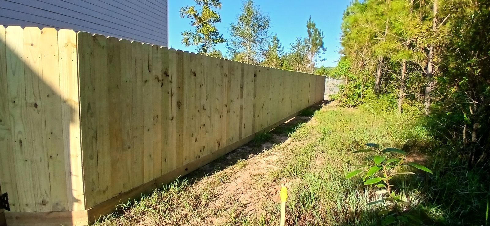 A wooden fence runs along a grassy area, separating a building from a tree line on a sunny day.