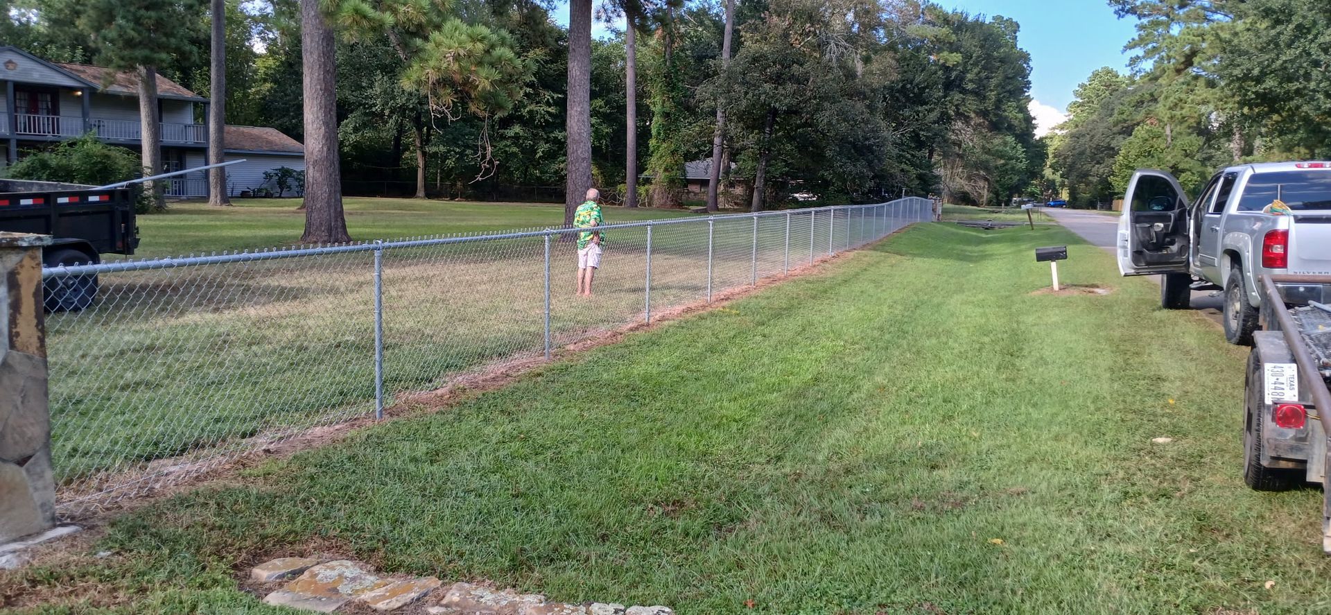 Chain-link fence borders a grassy area next to a road, with a truck and trailer parked on the right.