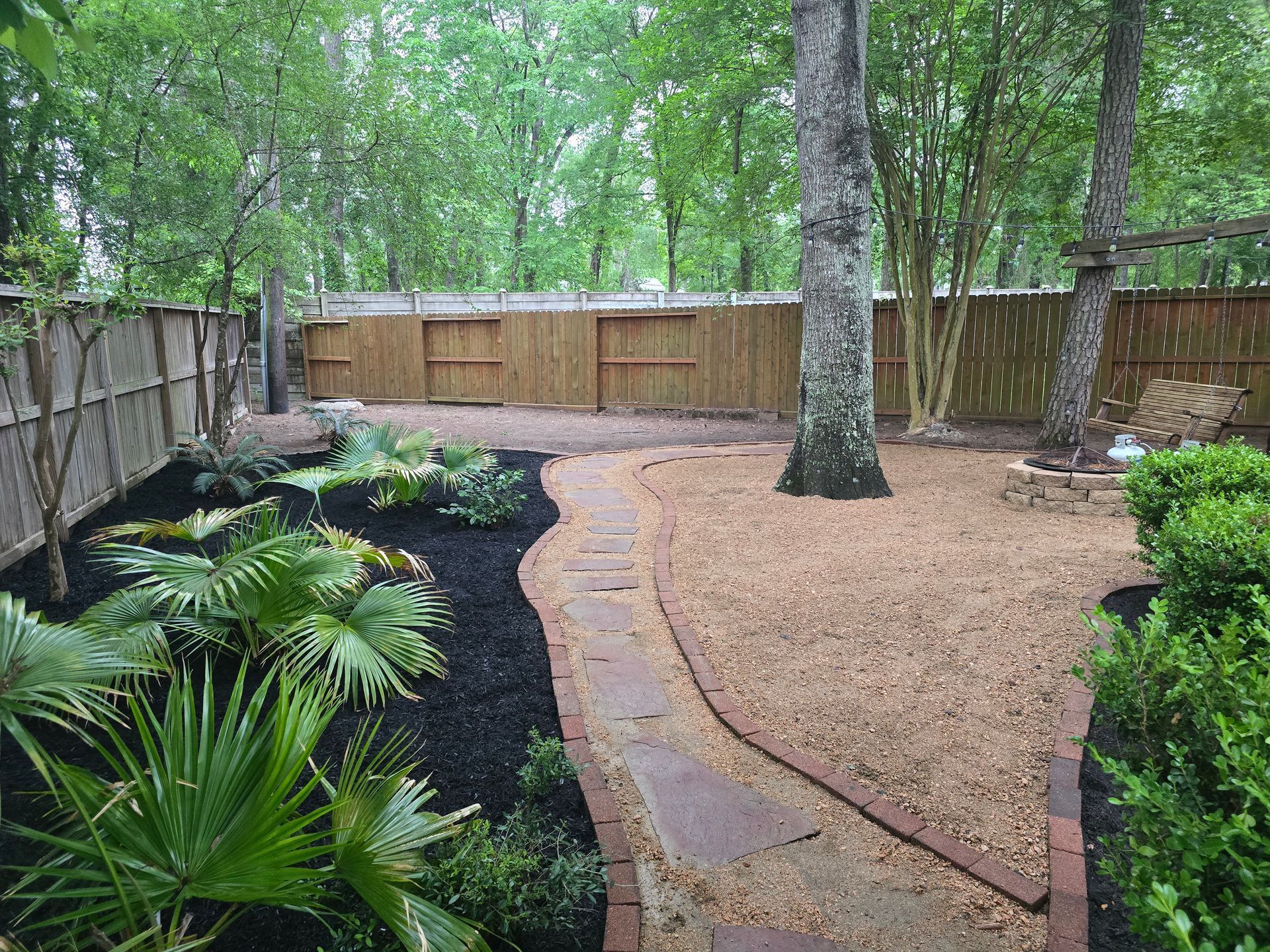 Backyard with landscaping, brick border pathway, mulch, trees, and wooden fence.