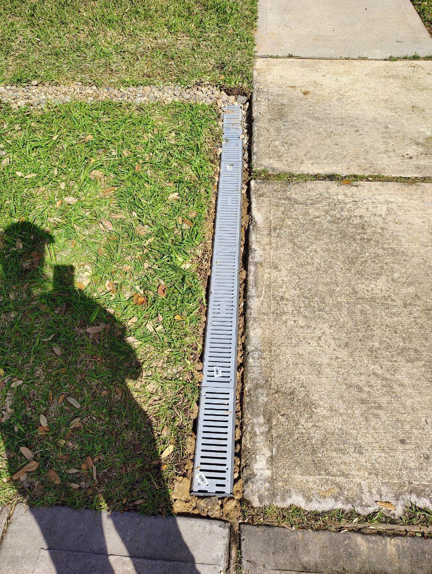 Grassy lawn and sidewalk separated by a long metal grate drain. Shadow of person on the lawn.