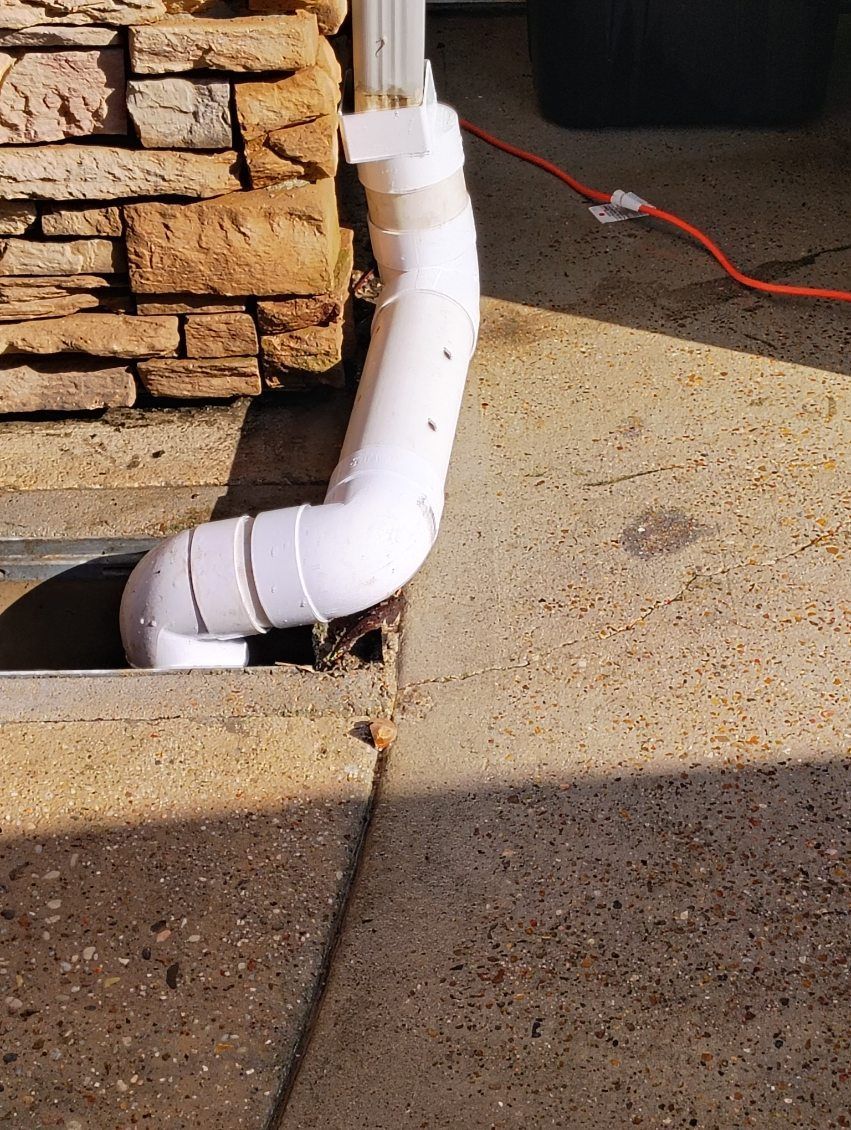 White PVC pipe diverts rainwater from a gutter down a wall and into a ground drain.