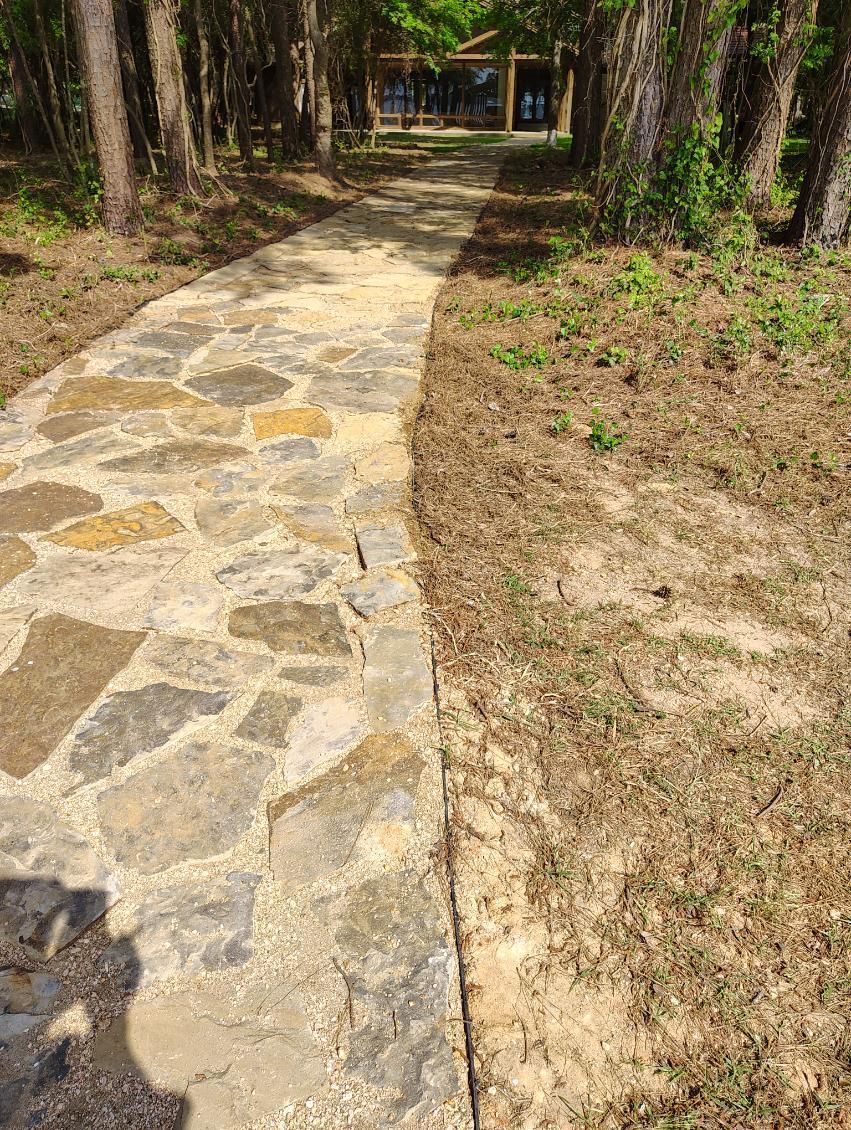 Stone path with grass on one side, trees in background leading to a structure.