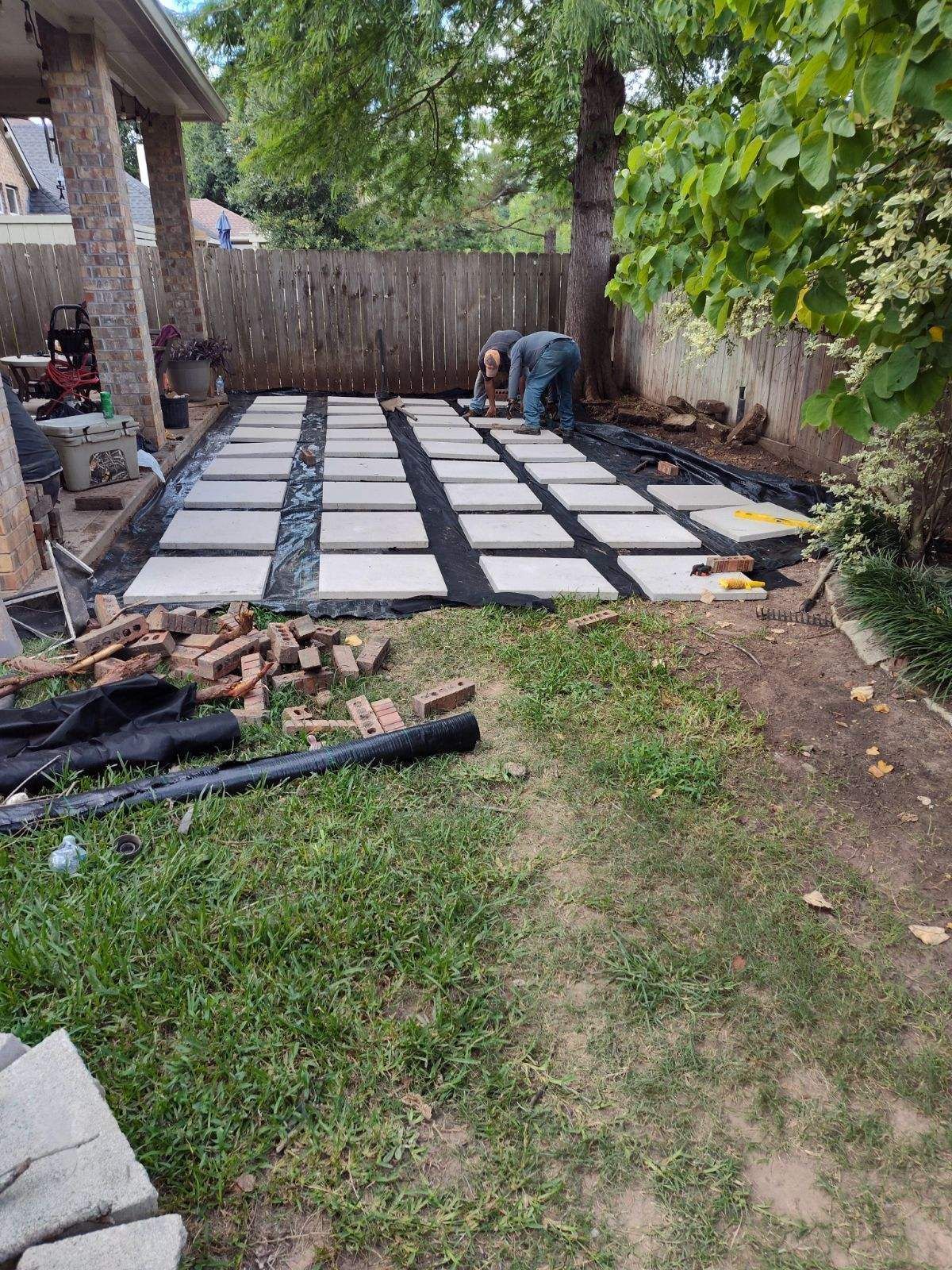 Construction of a patio with pavers laid on black fabric; a person works on the pavers.