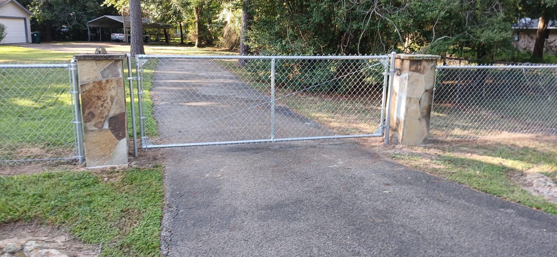 Chain link fence gate with stone pillars in front of a driveway. Trees and a house in the background.