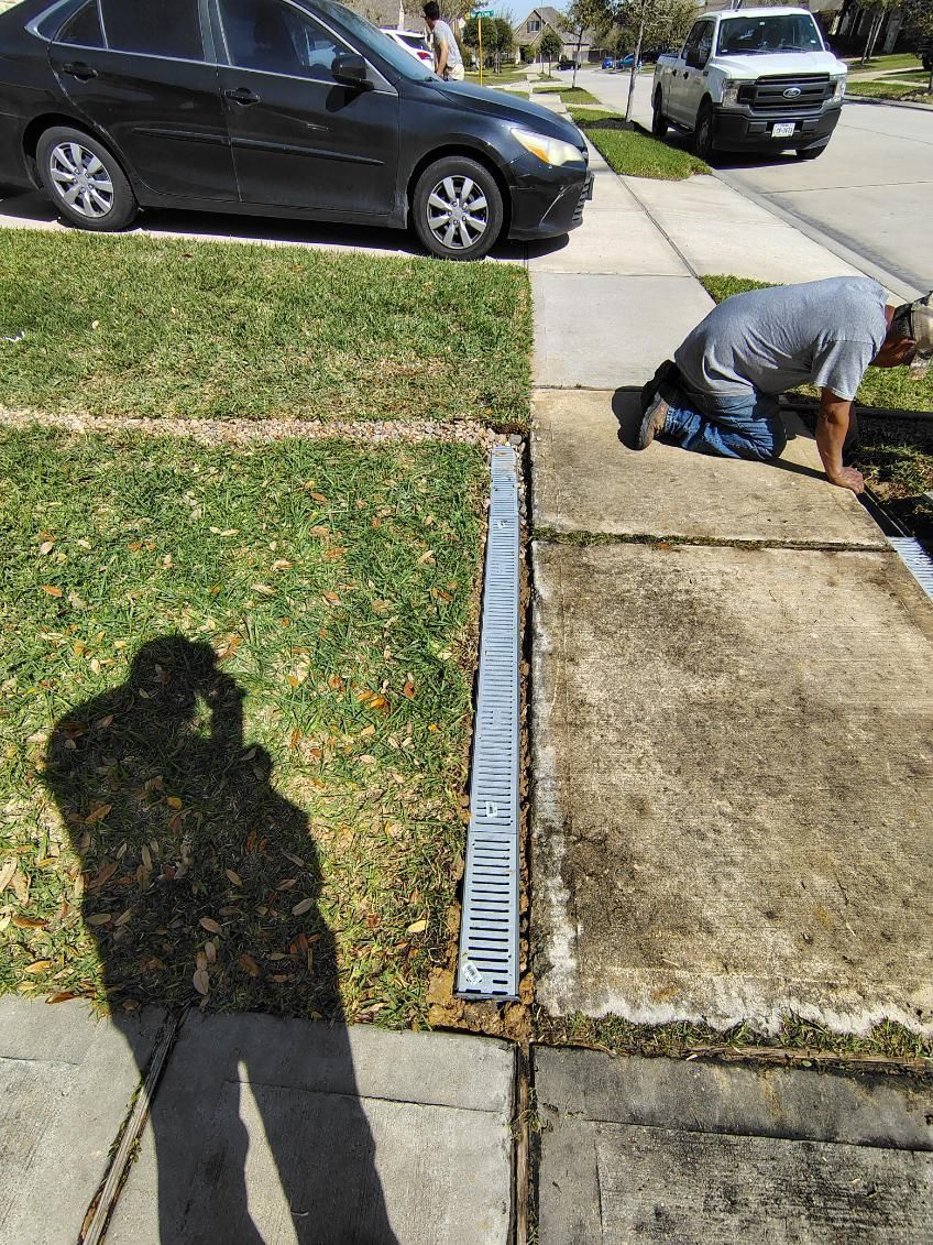 Man installing a metal drain along a sidewalk next to a lawn. Car parked nearby.
