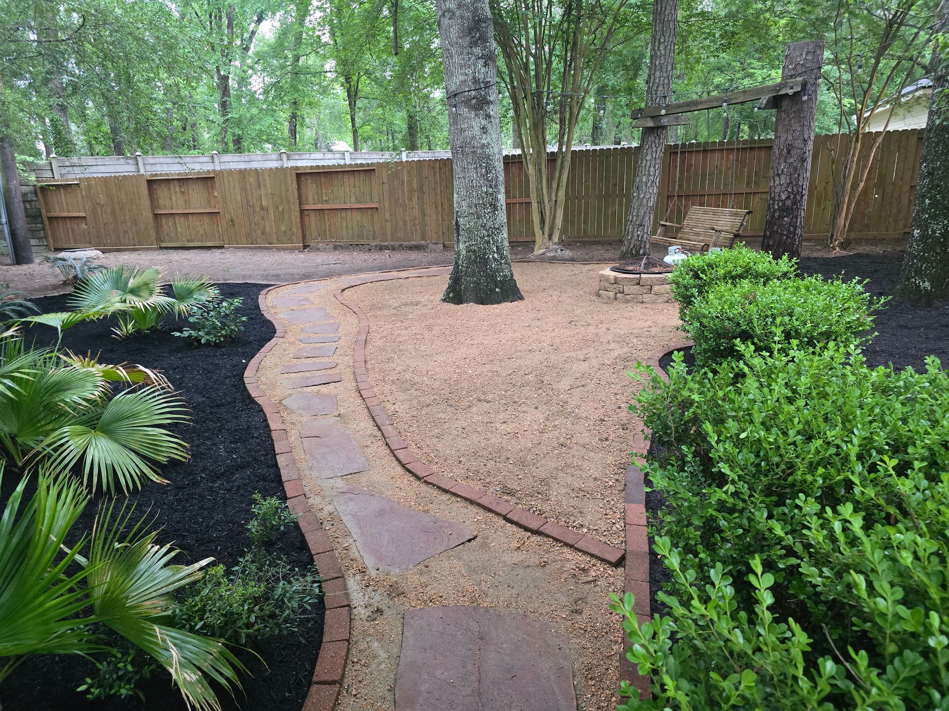 Backyard with stone path, brown gravel, mulch, and lush greenery. Wooden fence and trees in background.