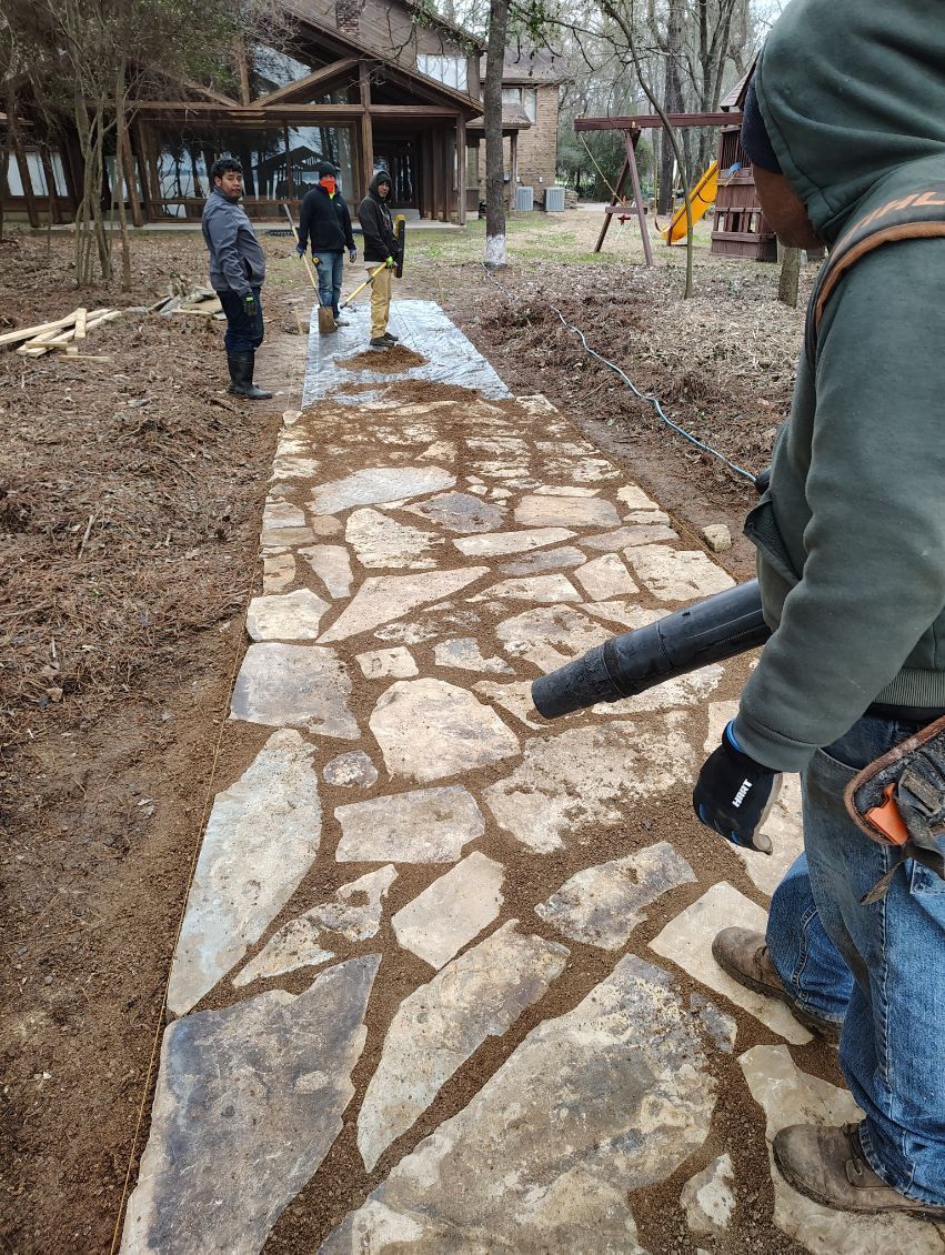 Man blowing debris off a stone pathway with two others in the background. A house is in the background.