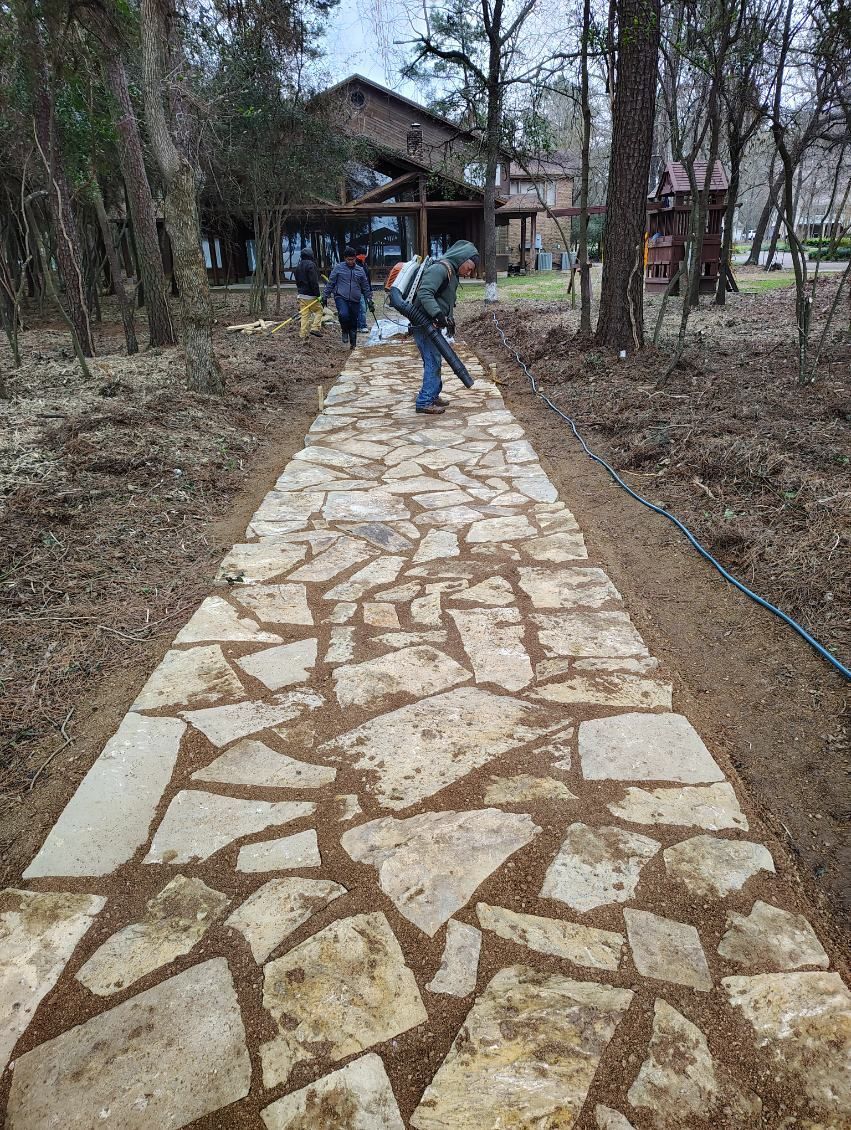 Stone pathway being cleared with a leaf blower, leading toward a building in a wooded area.