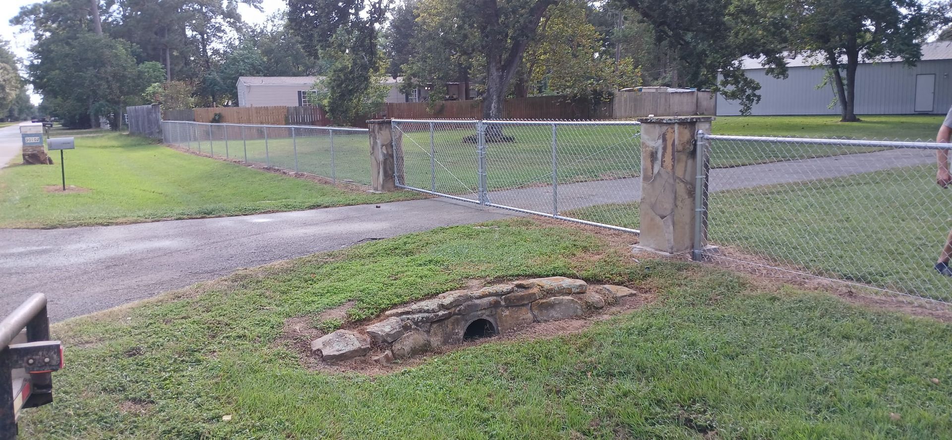 A chain-link fence gate with stone pillars in a grassy area, leading to a road. Trees in the background.