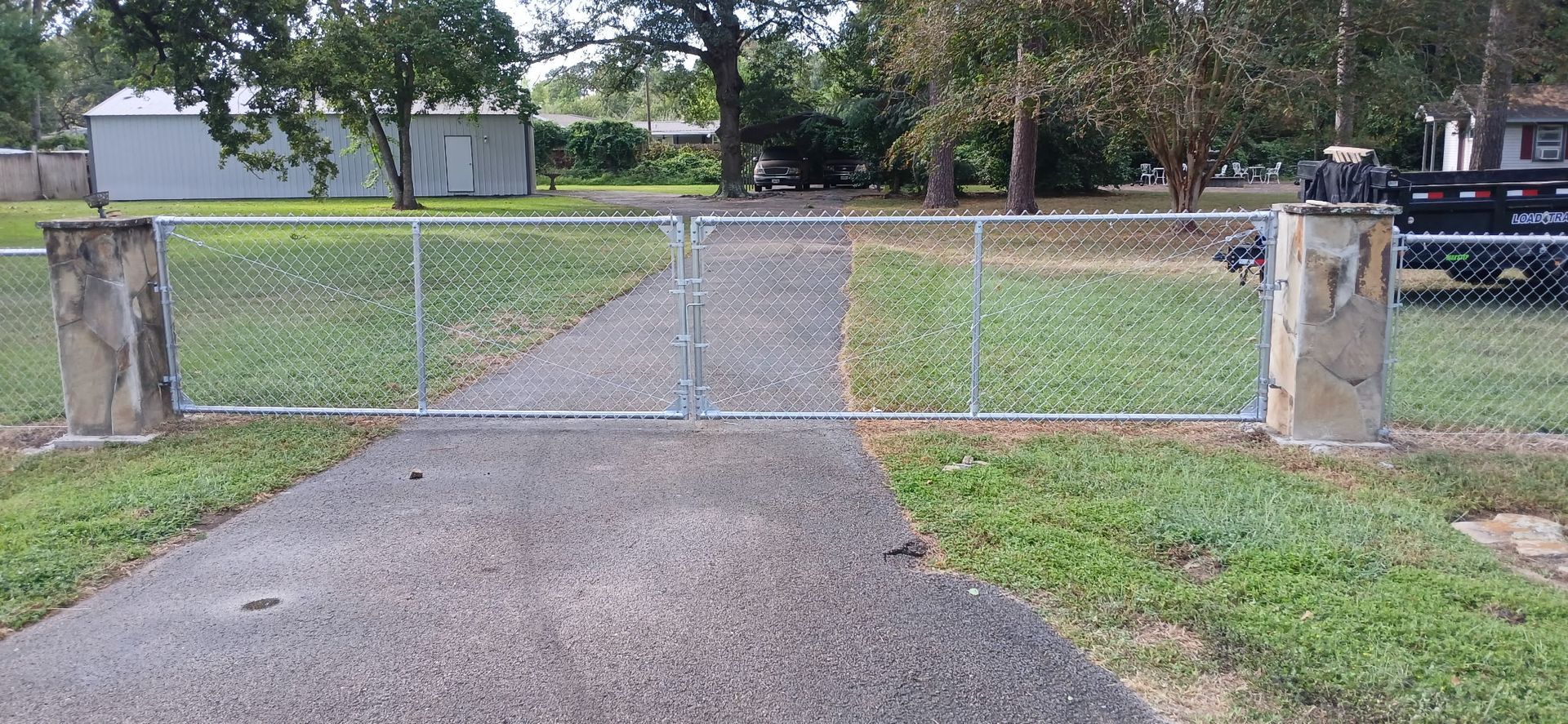 Chain-link gate between stone pillars, set in a paved driveway, surrounded by grass and trees.